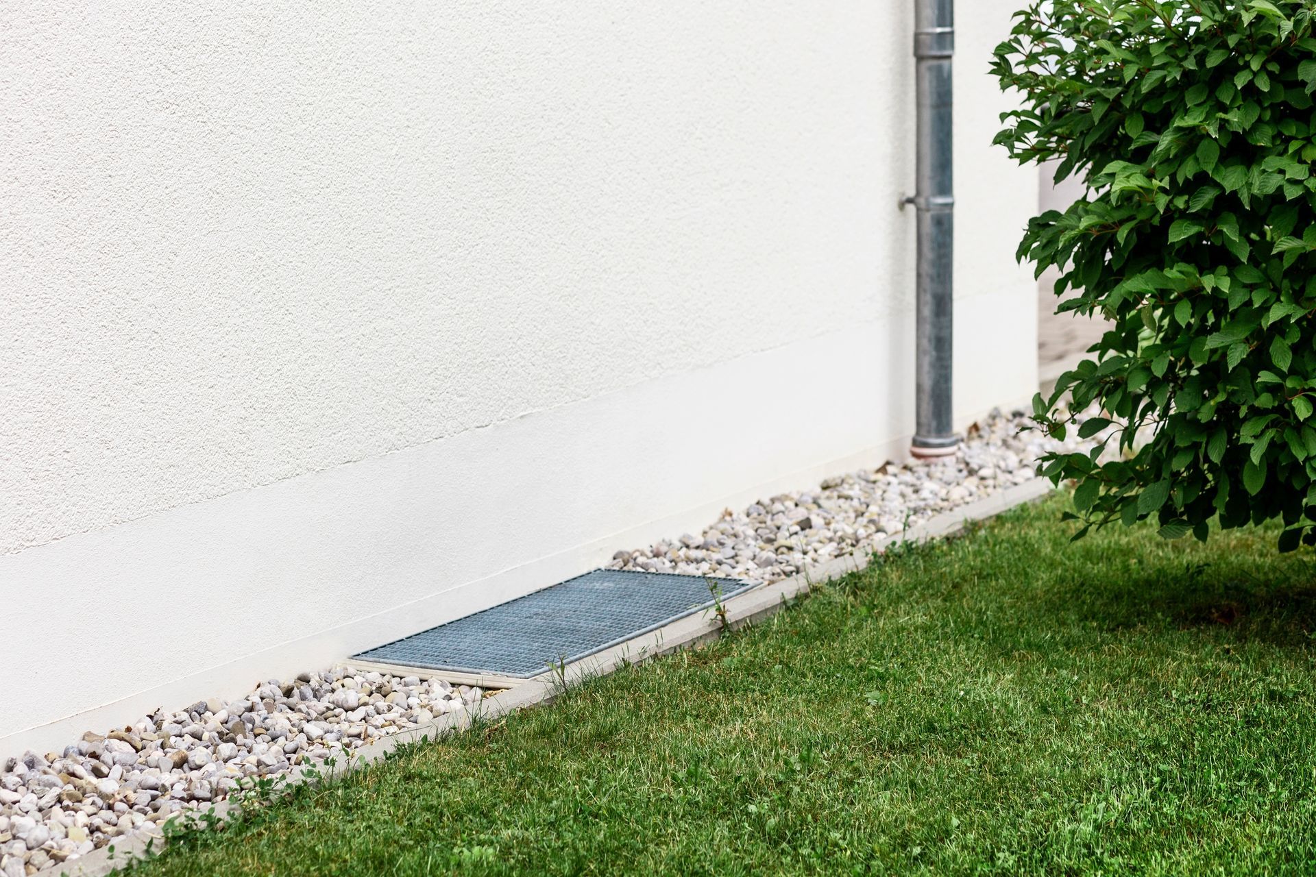White wall with a metal grate at the bottom, surrounded by gravel and grass. A downspout and a bush are to the right.