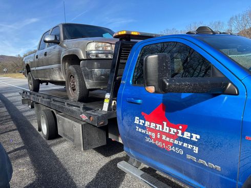 Blue tow truck hauling a gray pickup on a flatbed beside a snowy roadside