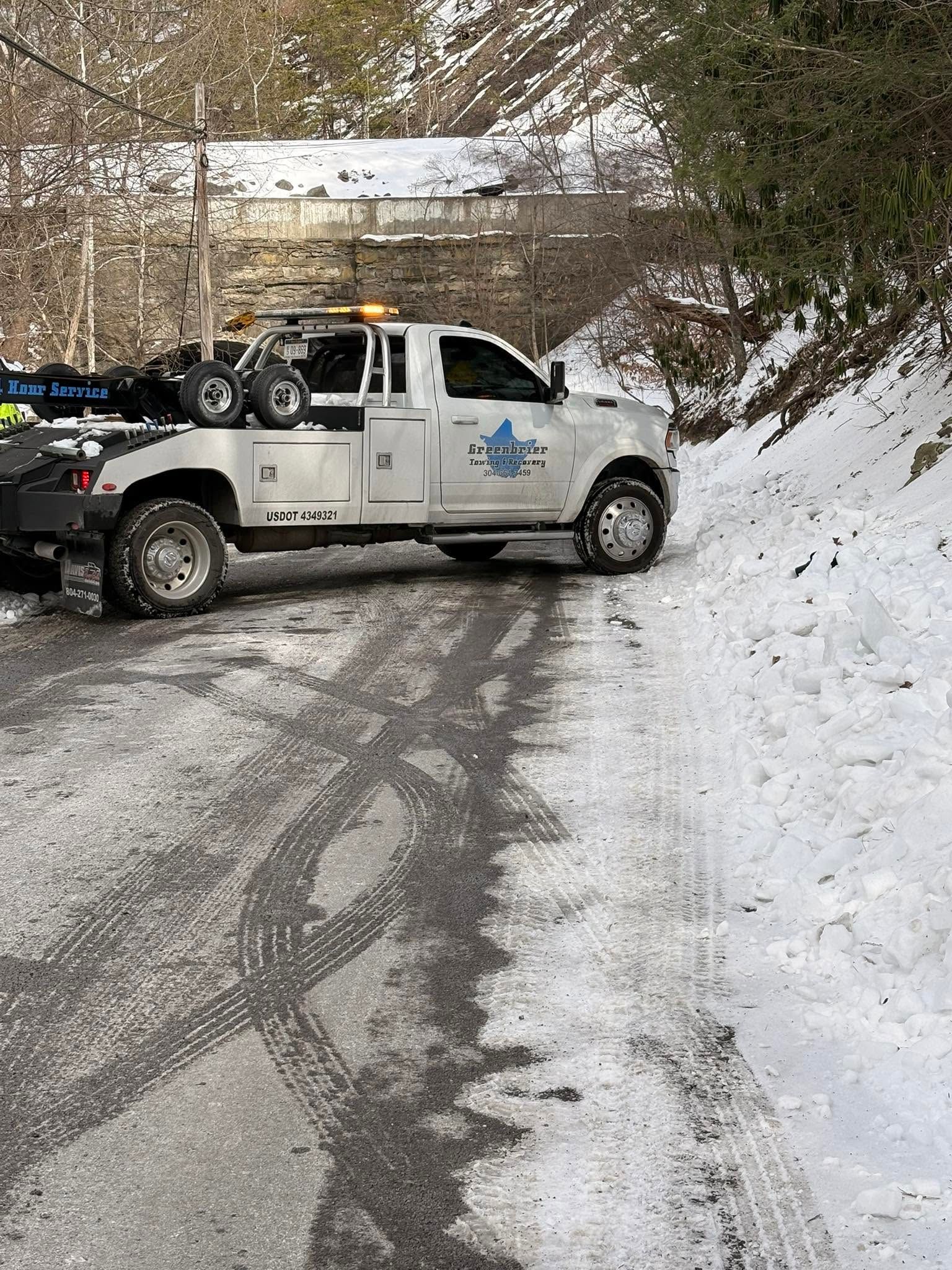 White tow truck on a snowy road beside a wooded hillside, towing a dark vehicle.