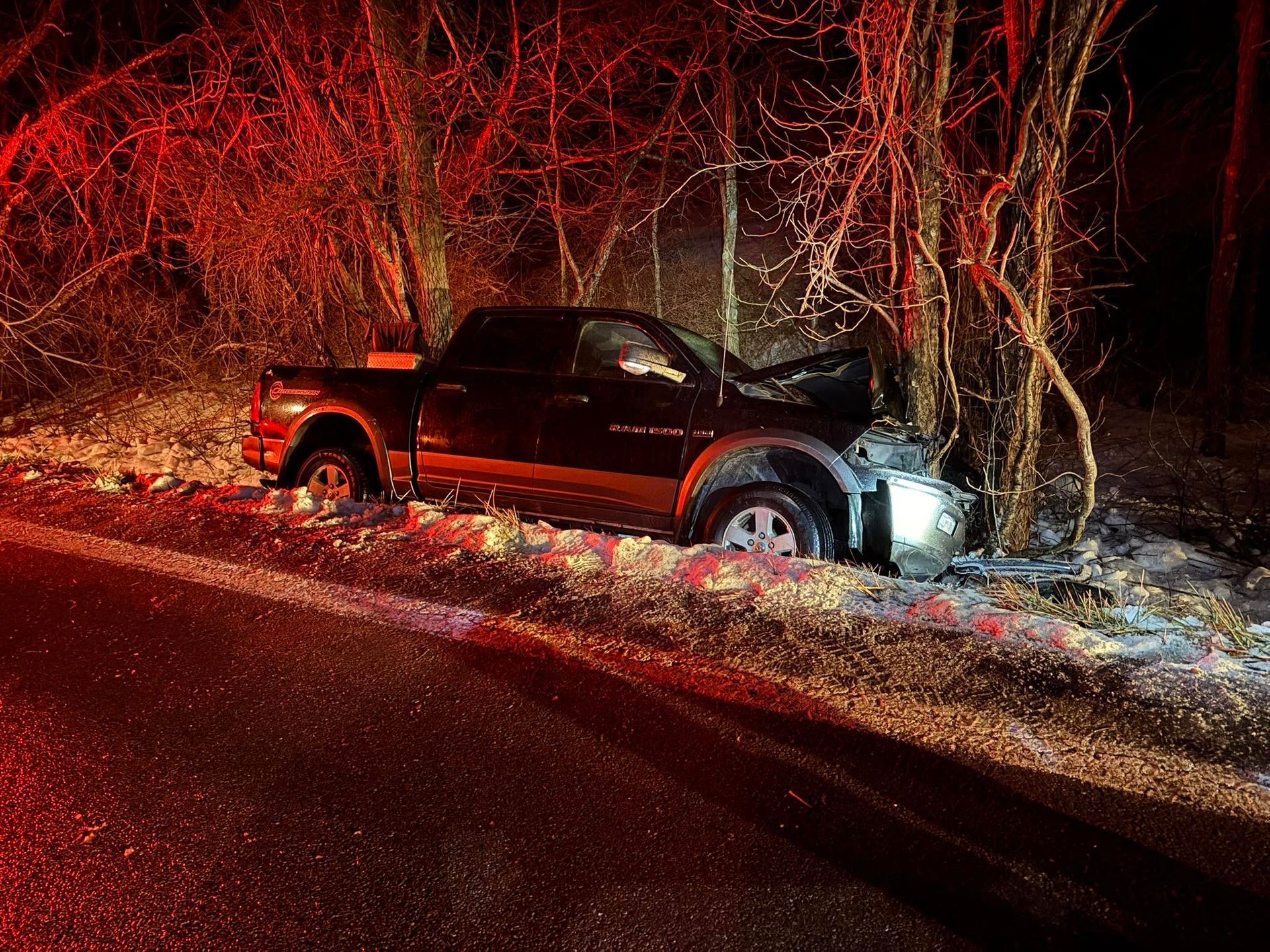 Black pickup truck wrecked in a ditch at night, headlights on, beside a snowy roadside and bare trees