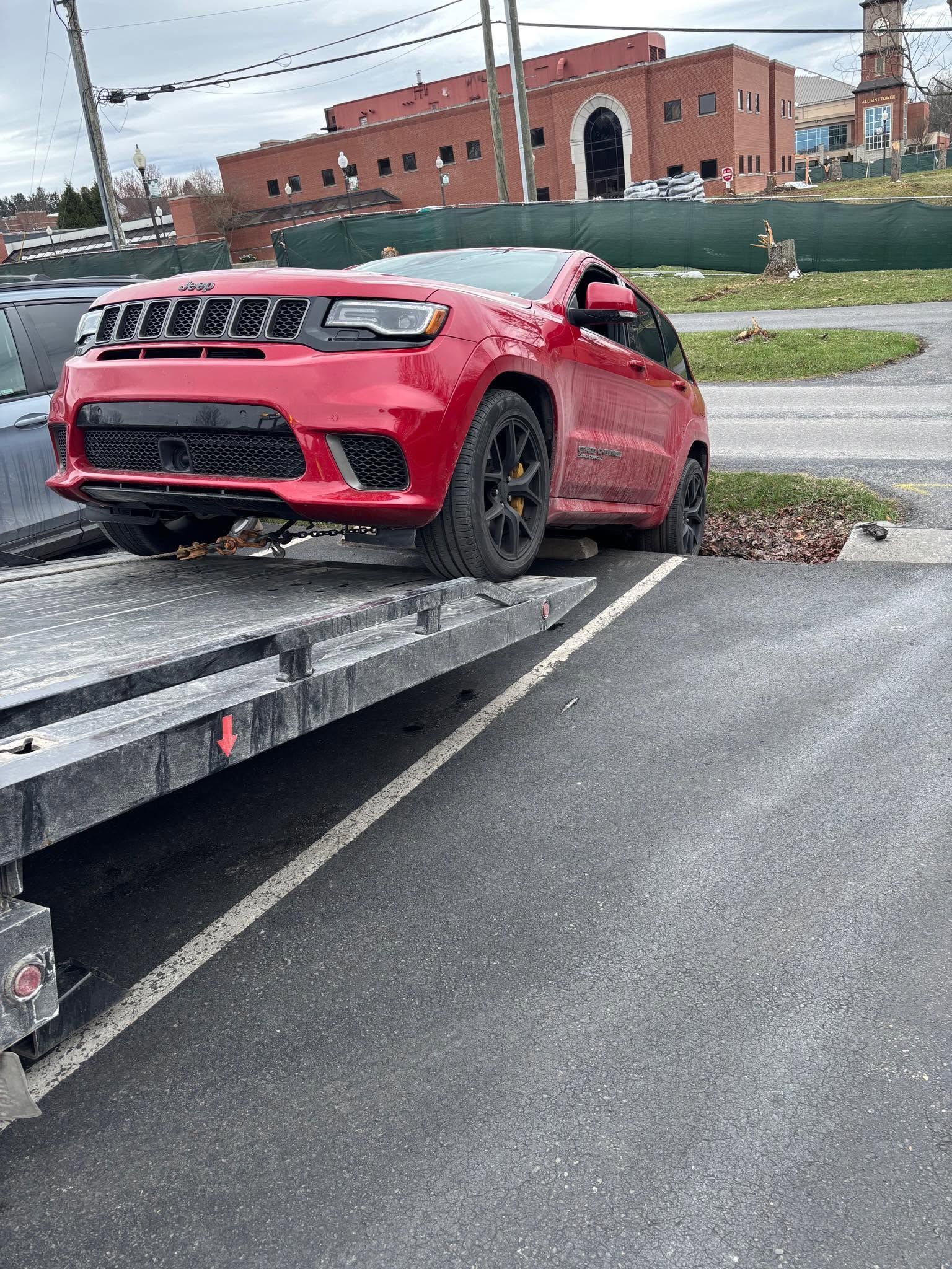 Red car partially off a flatbed tow truck on a wet roadside near a brick building.