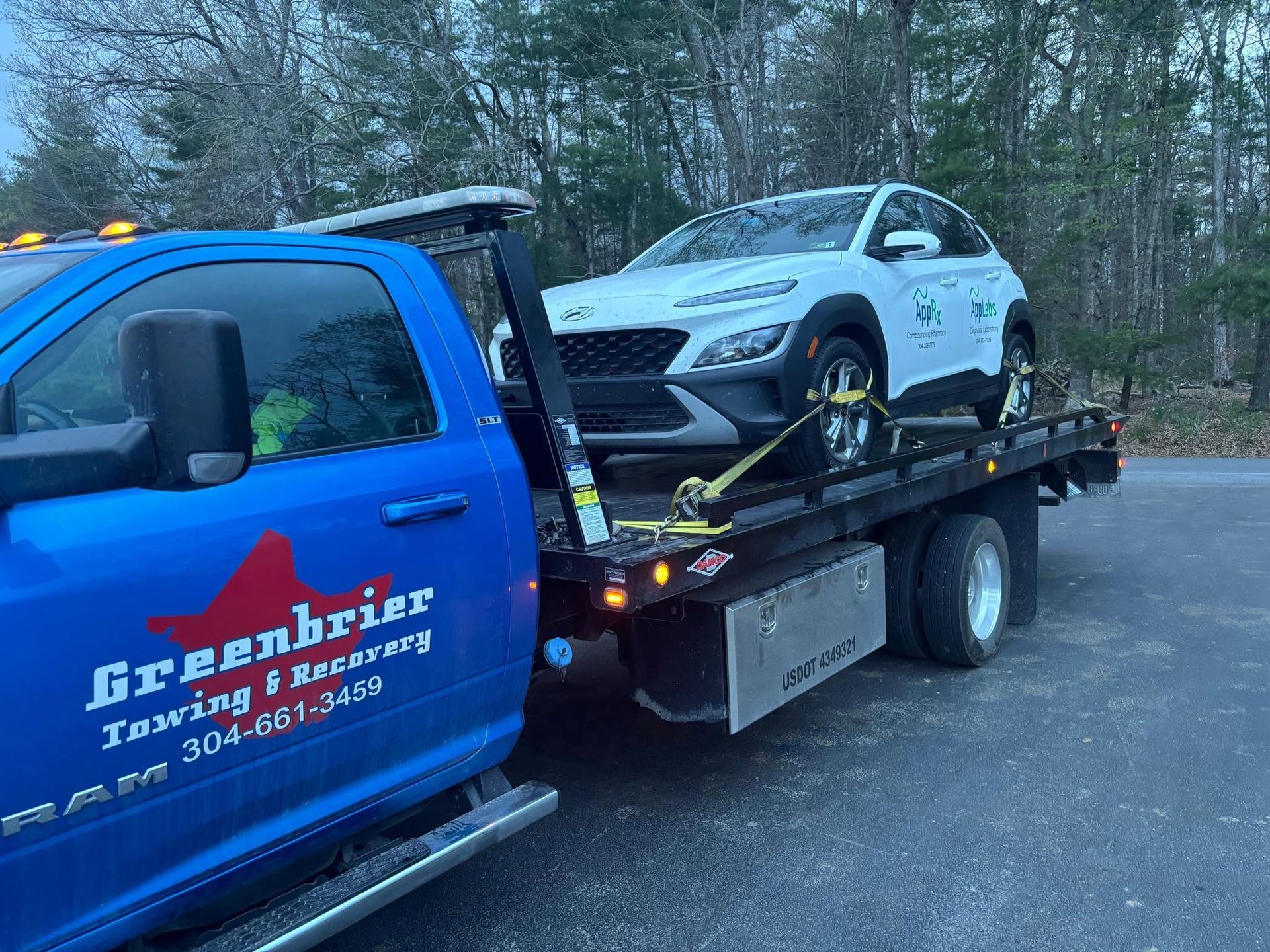 Blue tow truck hauling a white car on a flatbed beside a wooded roadside