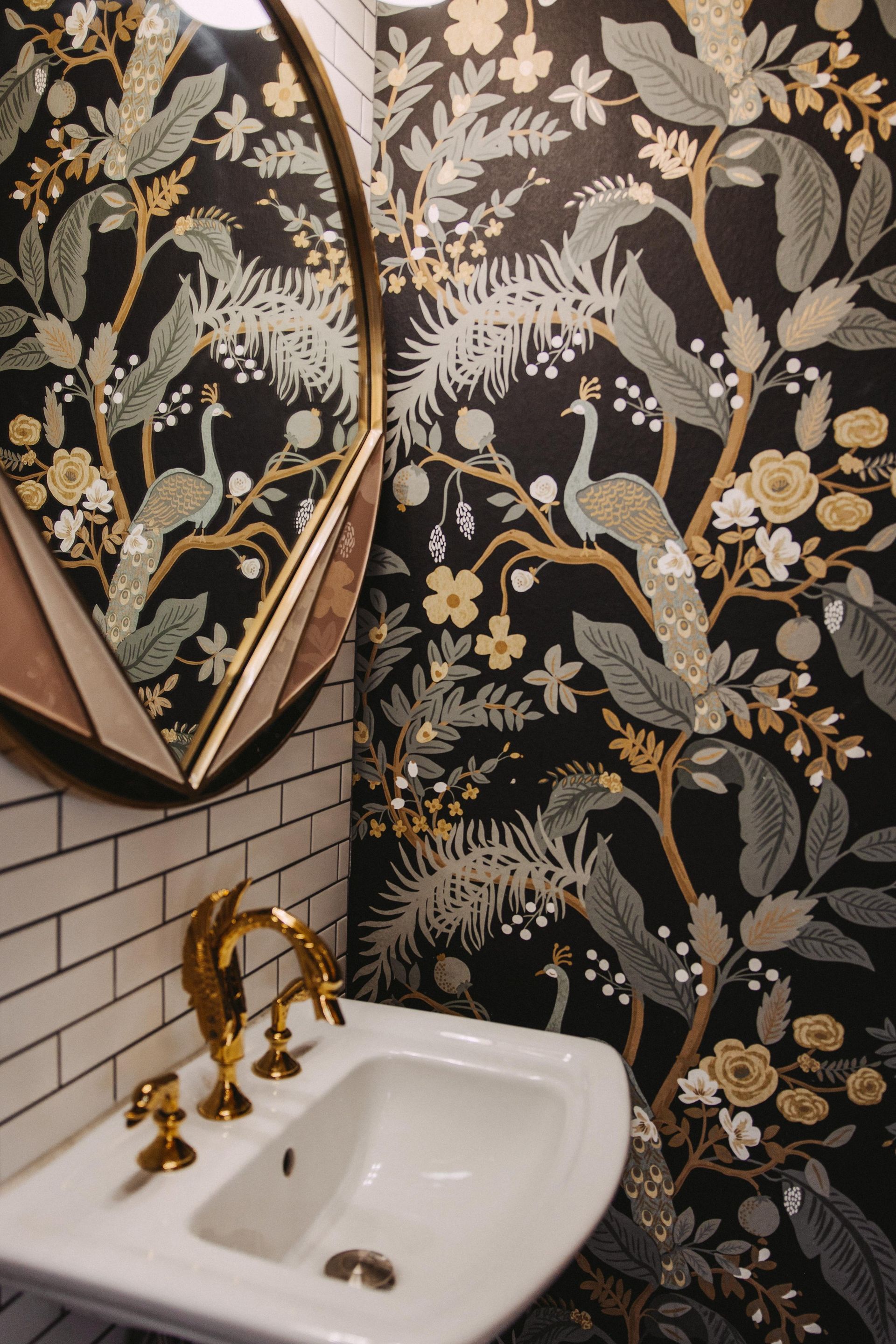 A stylish powder room with patterned wallpaper featuring birds and gold accents. It has a white sink with a gold faucet and a geometric mirror.