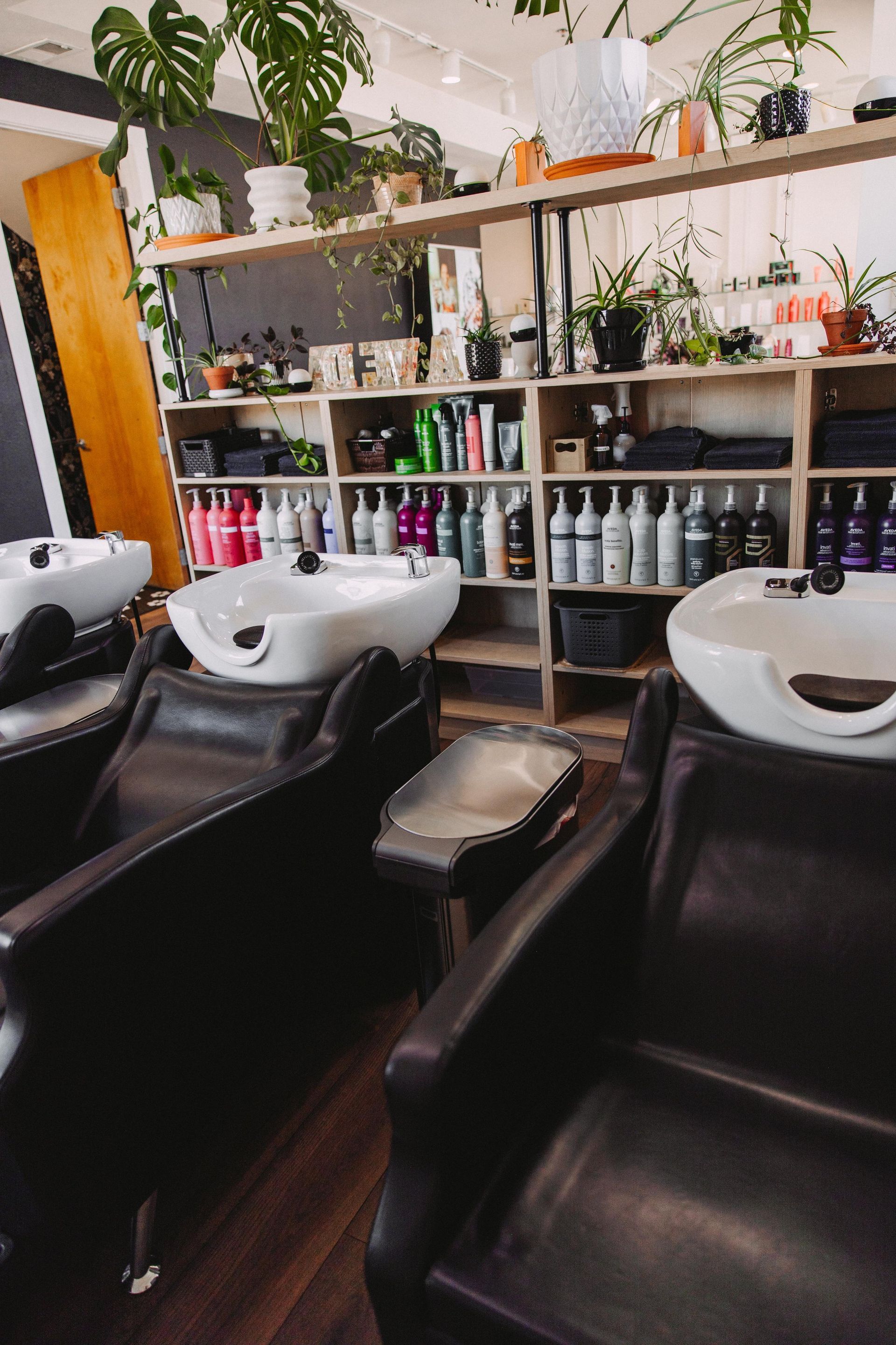 Salon interior with three wash basins, dark leather chairs, and shelves stocked with hair products.