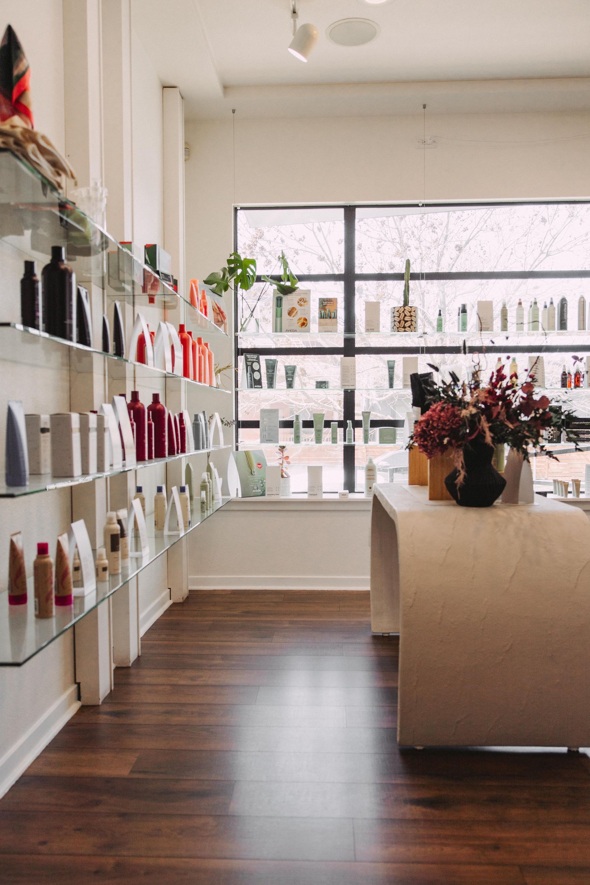 A well-lit beauty salon interior with products displayed on shelves, a large window, and a counter with flowers.