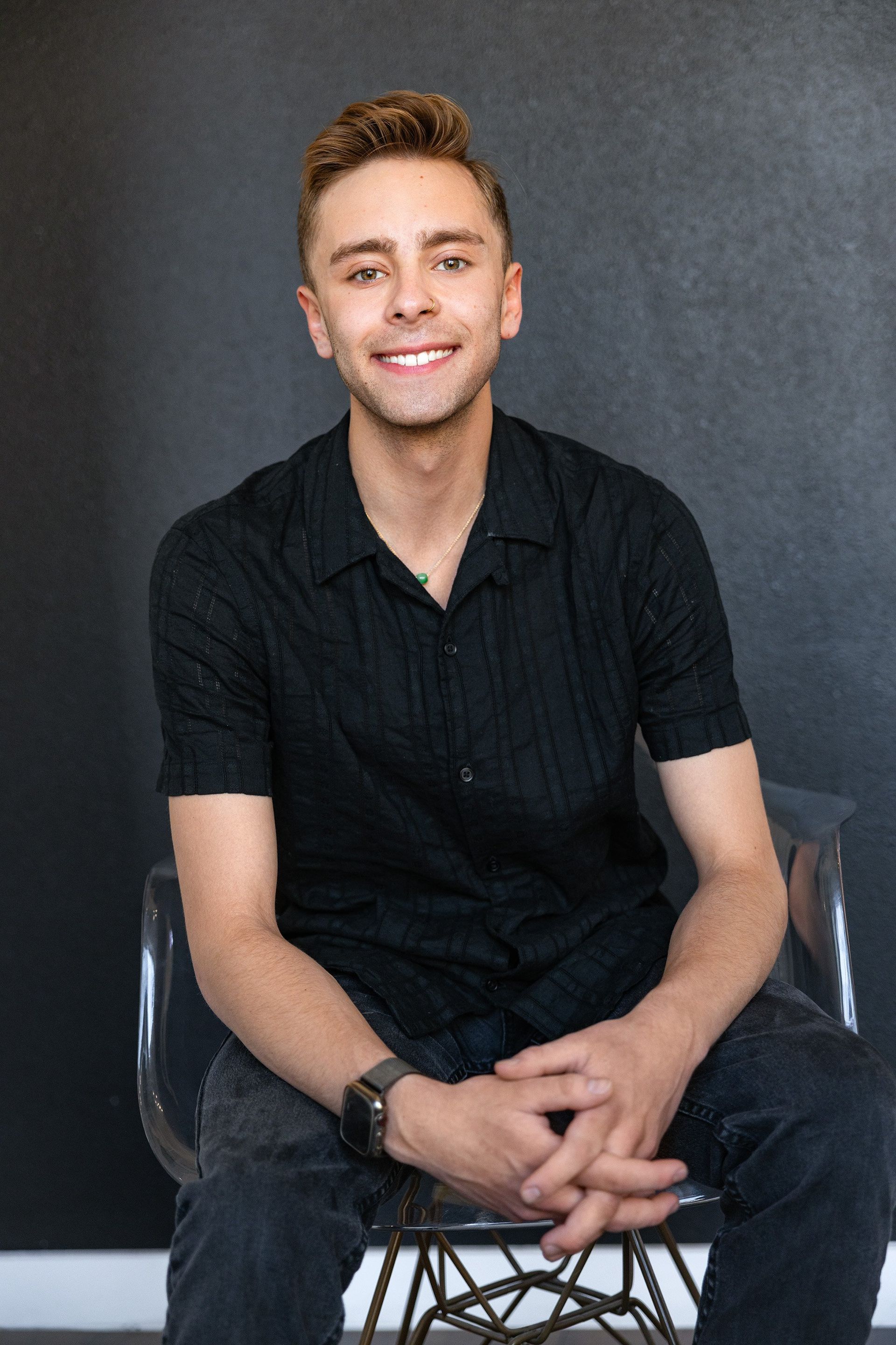 Young man smiles while sitting in a clear chair in front of a dark wall, wearing a black shirt and gray pants.