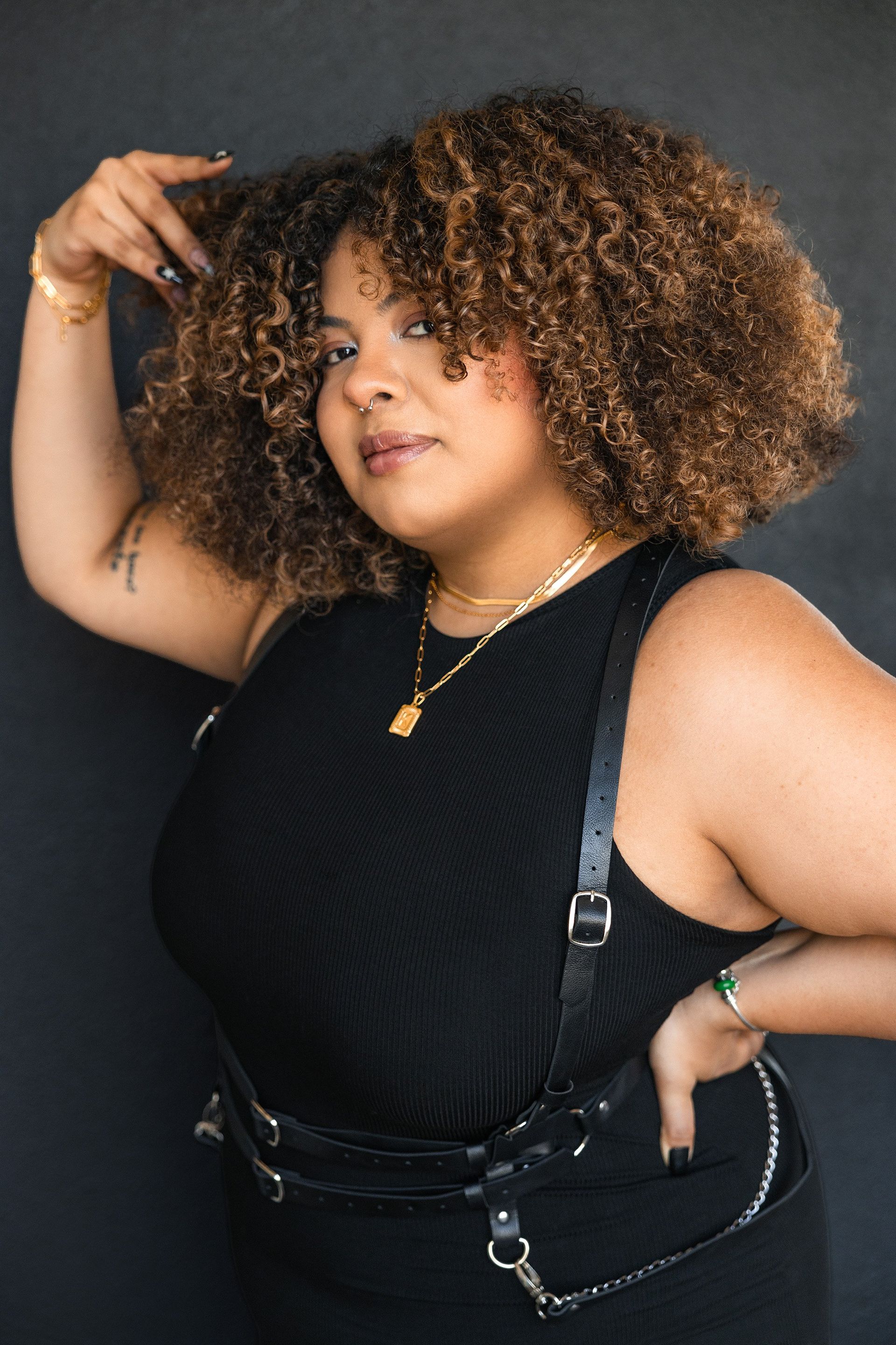 Woman with curly brown hair, wearing a black top, leather harness, and gold jewelry poses against a dark background.