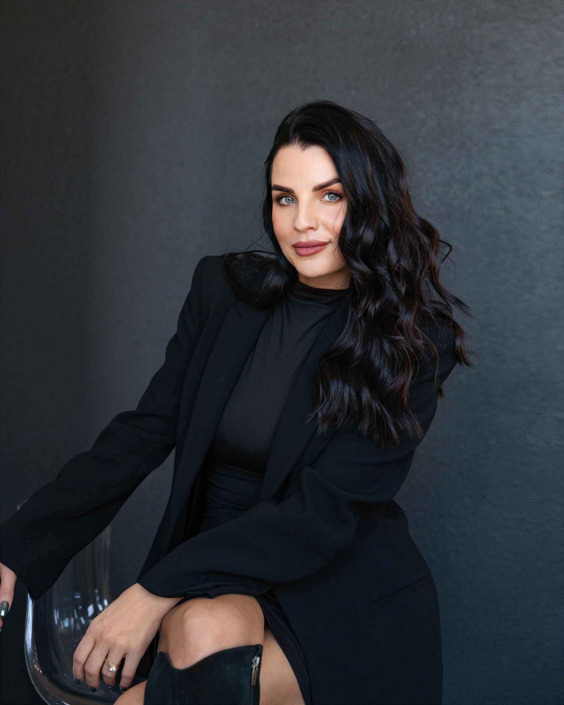 Woman with dark hair in a black blazer, top, and thigh-high boots, seated against a dark gray background.