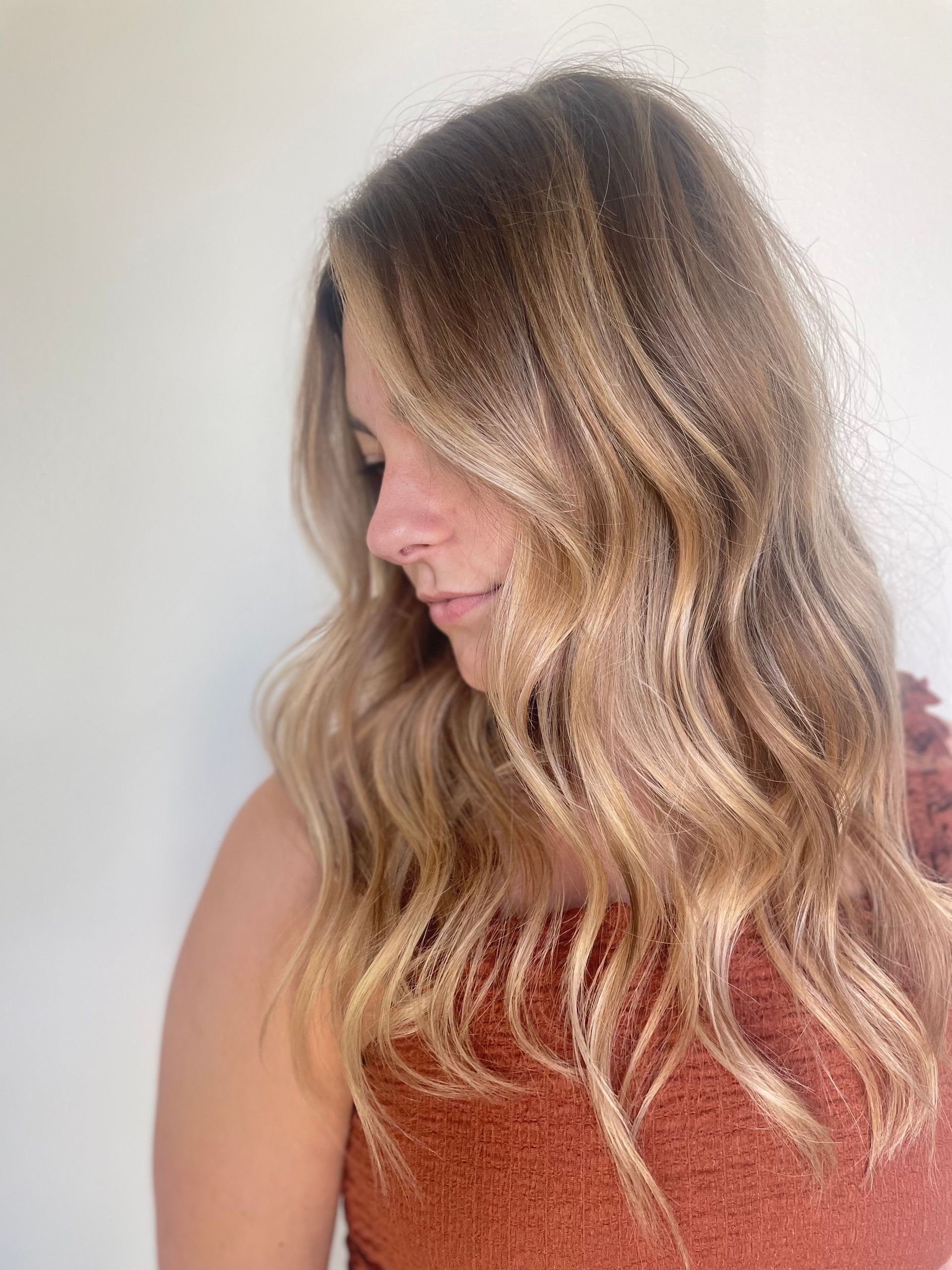 Woman with wavy blonde hair, side profile, looking down. Wearing a rust-colored top against a white background.