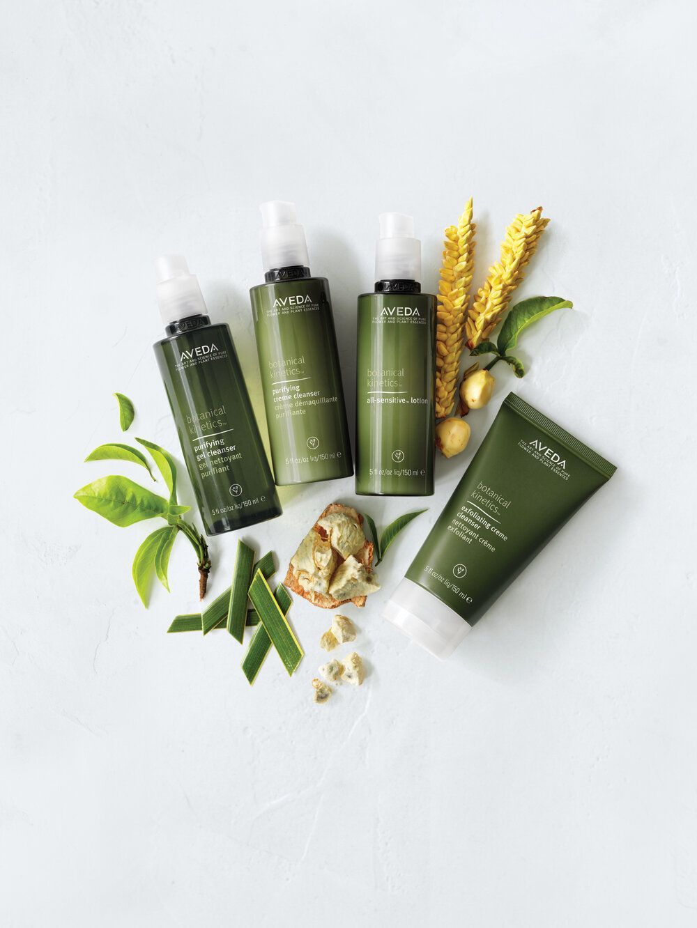 Green skincare products with botanicals on a white background. Bottles and a tube are arranged with leaves, plants, and roots.