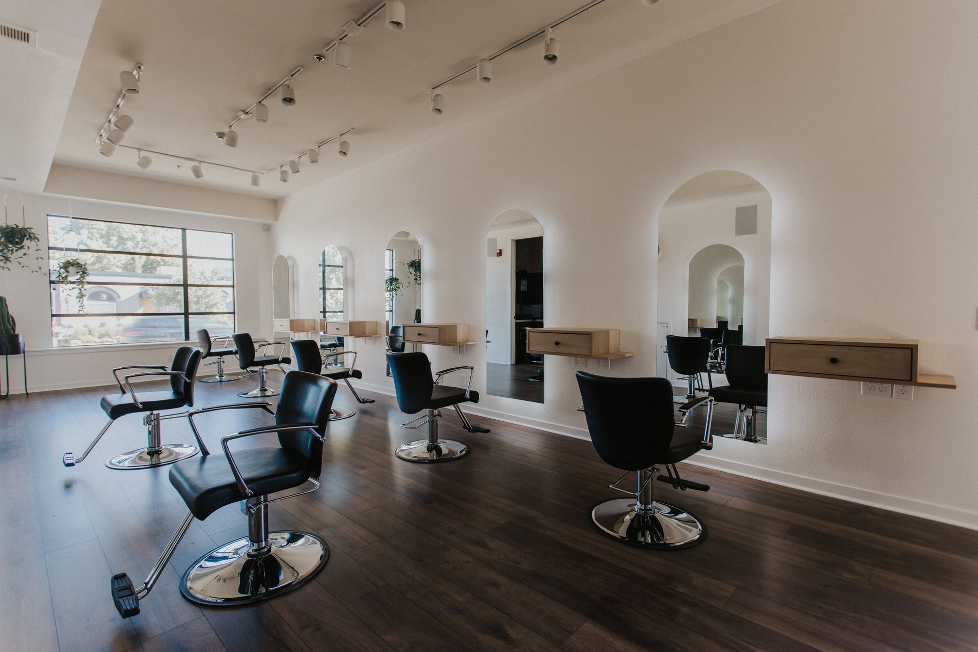 A modern hair salon with black chairs, large mirrors, and dark wood floors. Natural light streams in from a large window.