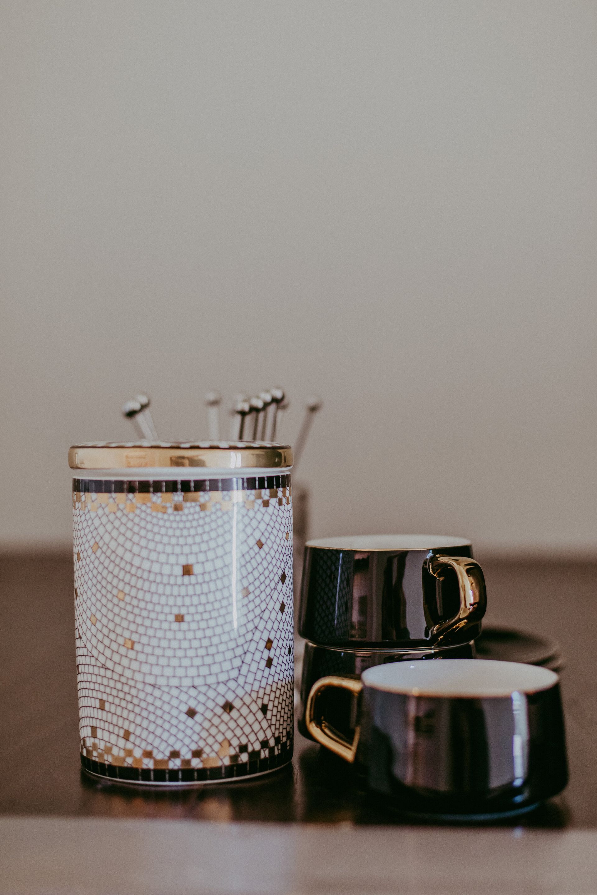 A cylindrical container with mosaic tile detail and two dark ceramic cups with gold handles sit on a table.