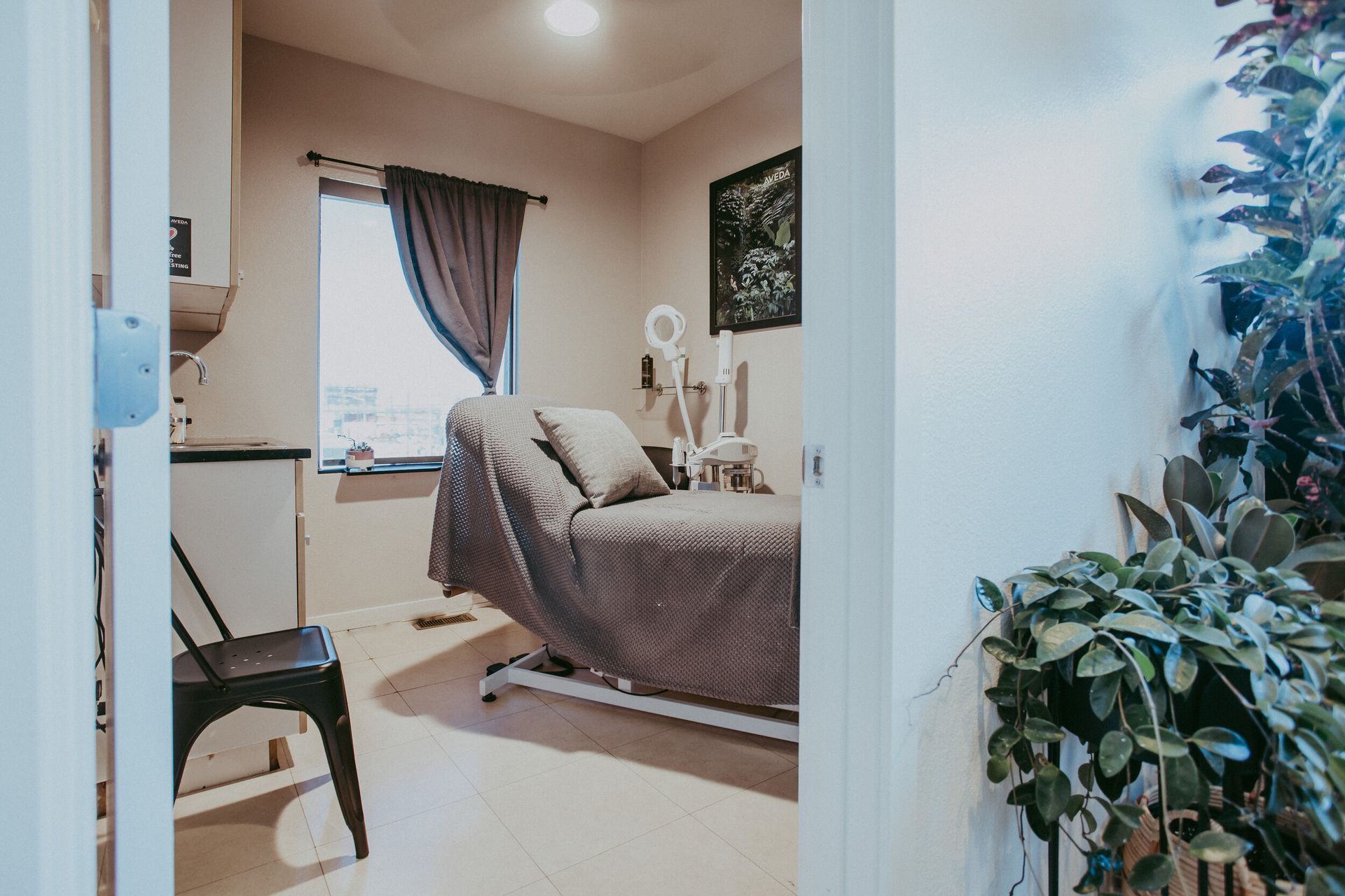 A room with a treatment bed, window, and plants seen through a doorway. The room has neutral colors and a relaxed atmosphere.