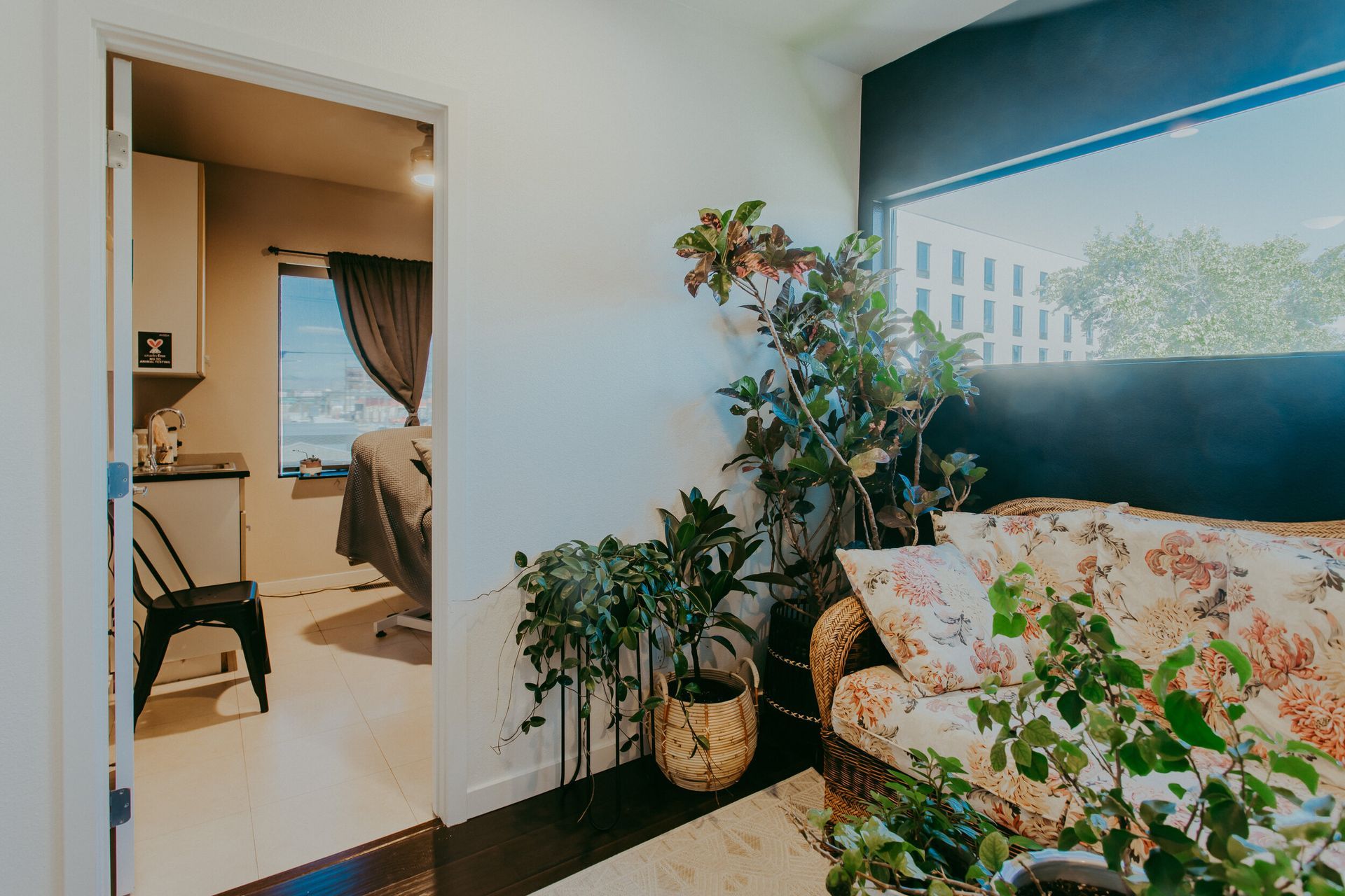 An interior view shows a floral patterned couch and plants near a window. An open doorway leads to another room with a desk.