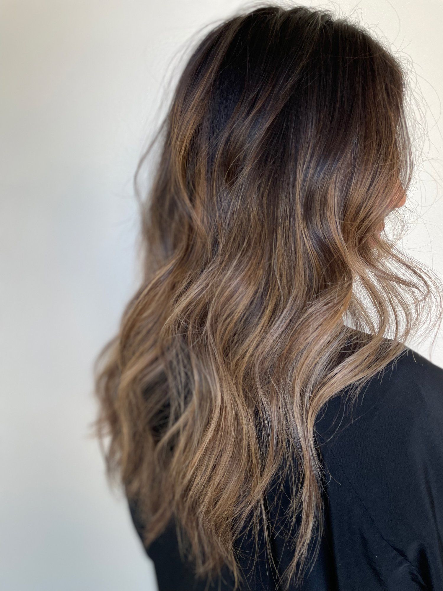 Woman with long, wavy brown hair featuring balayage highlights against a plain wall.