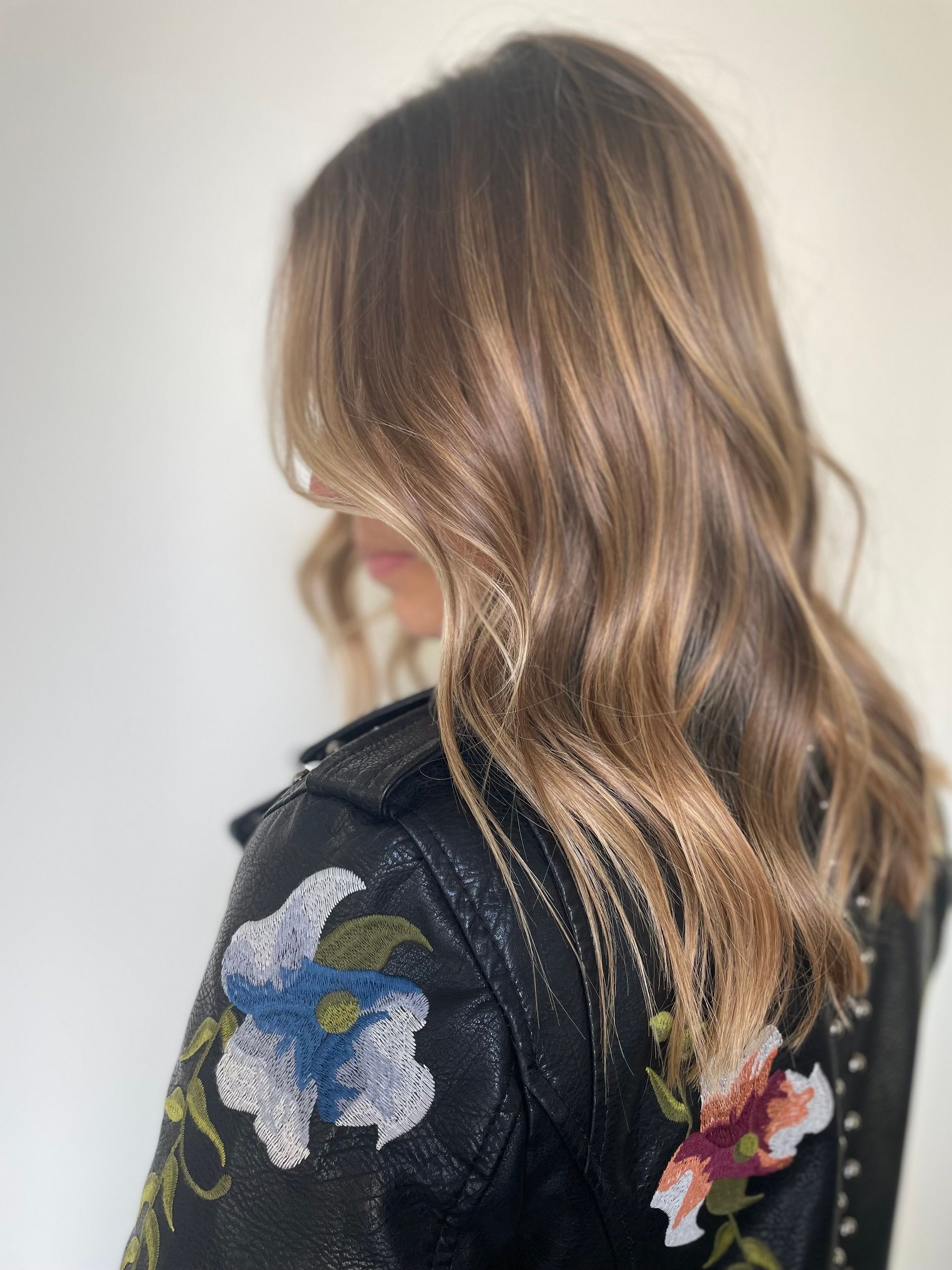 Woman with shoulder-length wavy brown hair and highlights, wearing a black jacket with floral embroidery; facing away from the camera.