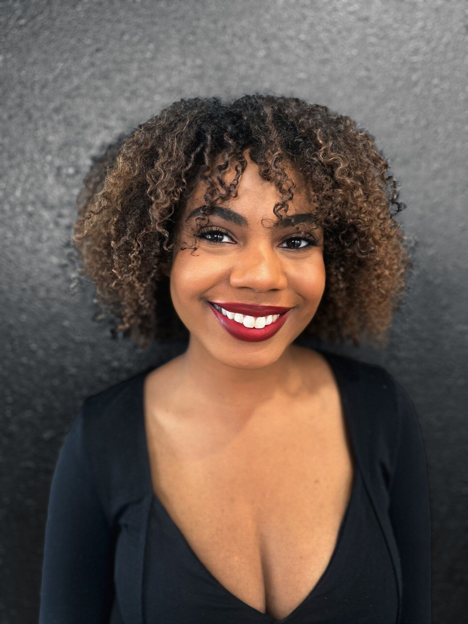 Smiling Black woman with curly hair and red lipstick, wearing a black top against a dark backdrop.