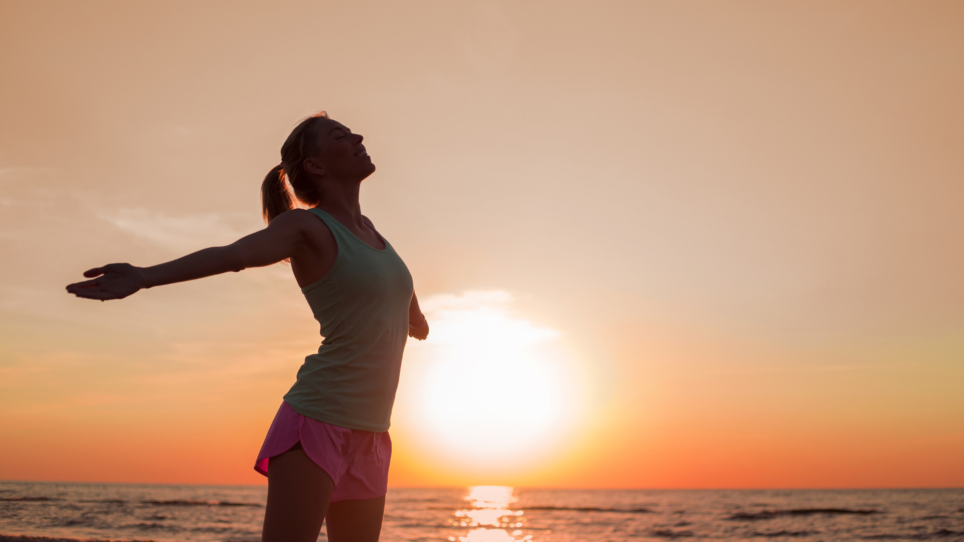 A woman is standing on the beach with her arms outstretched at sunset.