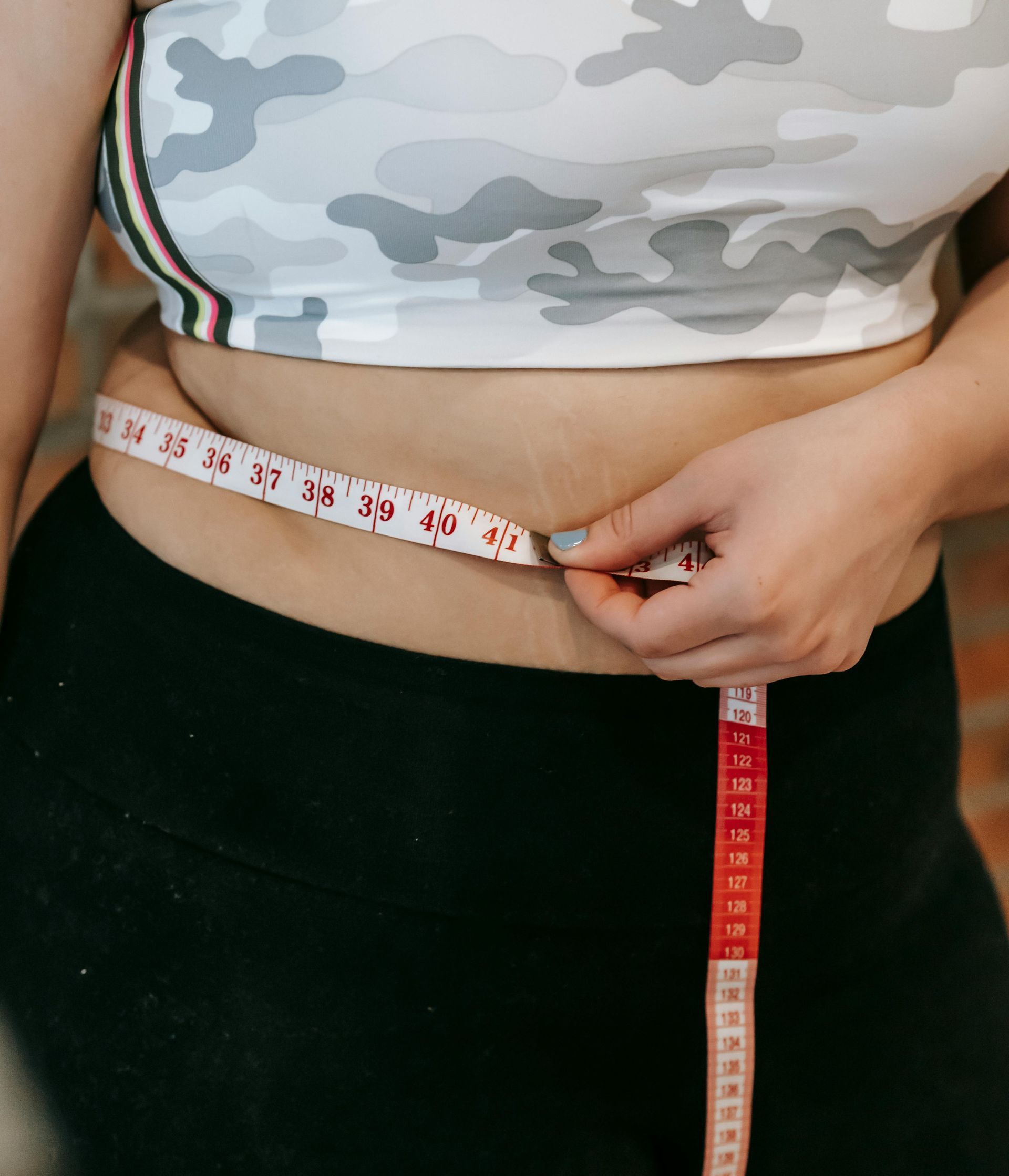 Person measuring waist with a tape measure. Wearing a camouflage crop top and black pants.