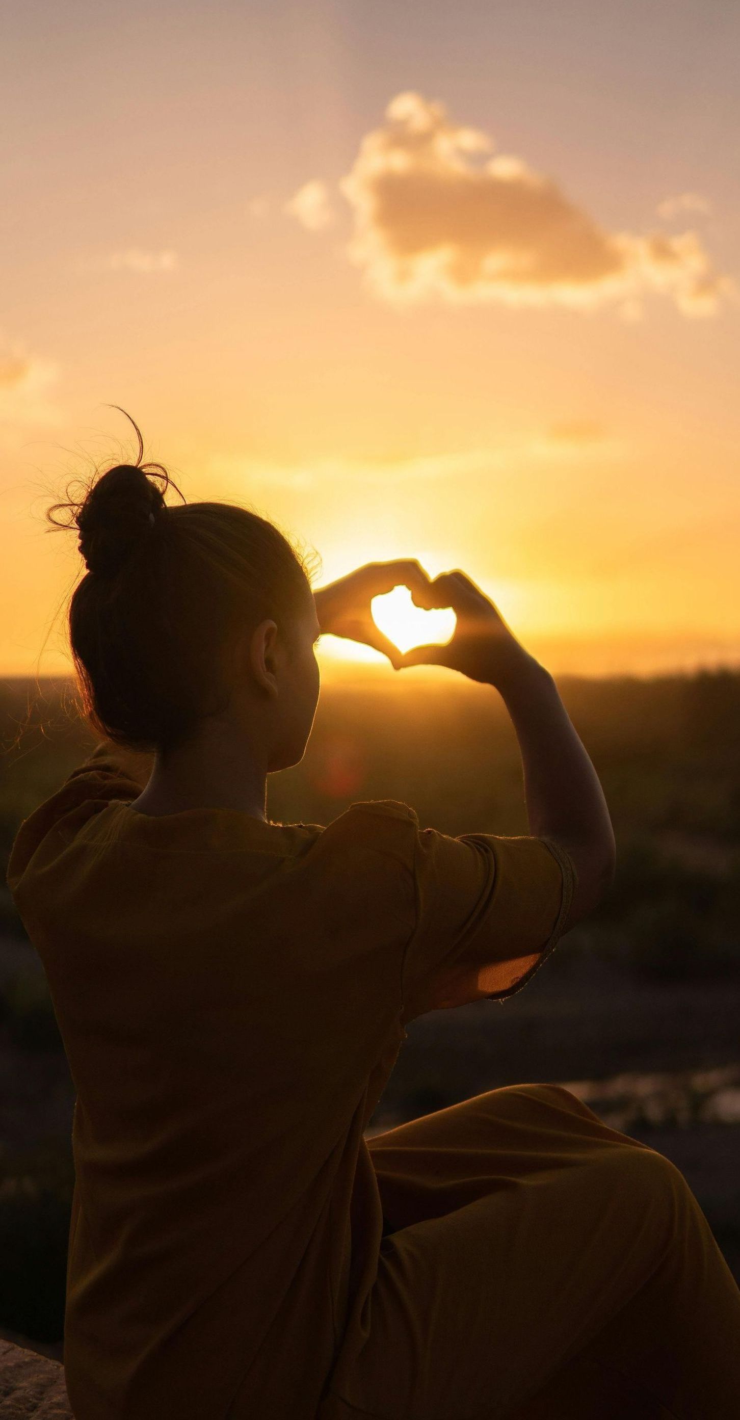A woman is making a heart shape with her hands at sunset.