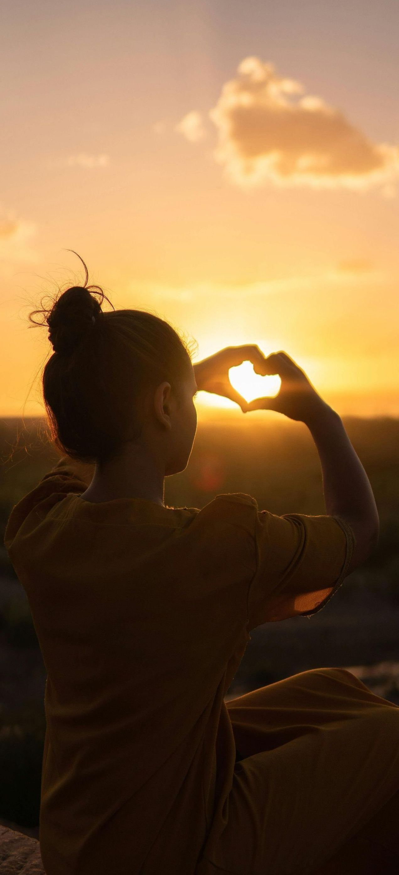 A woman is making a heart shape with her hands at sunset.