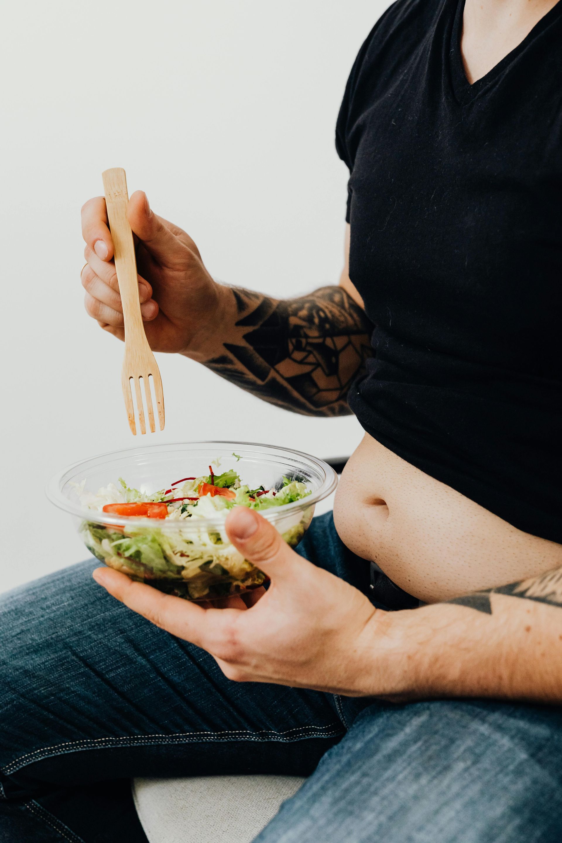 Person with tattoos eating salad from a clear bowl, wearing a black shirt and jeans. The person is sitting with a visible belly.