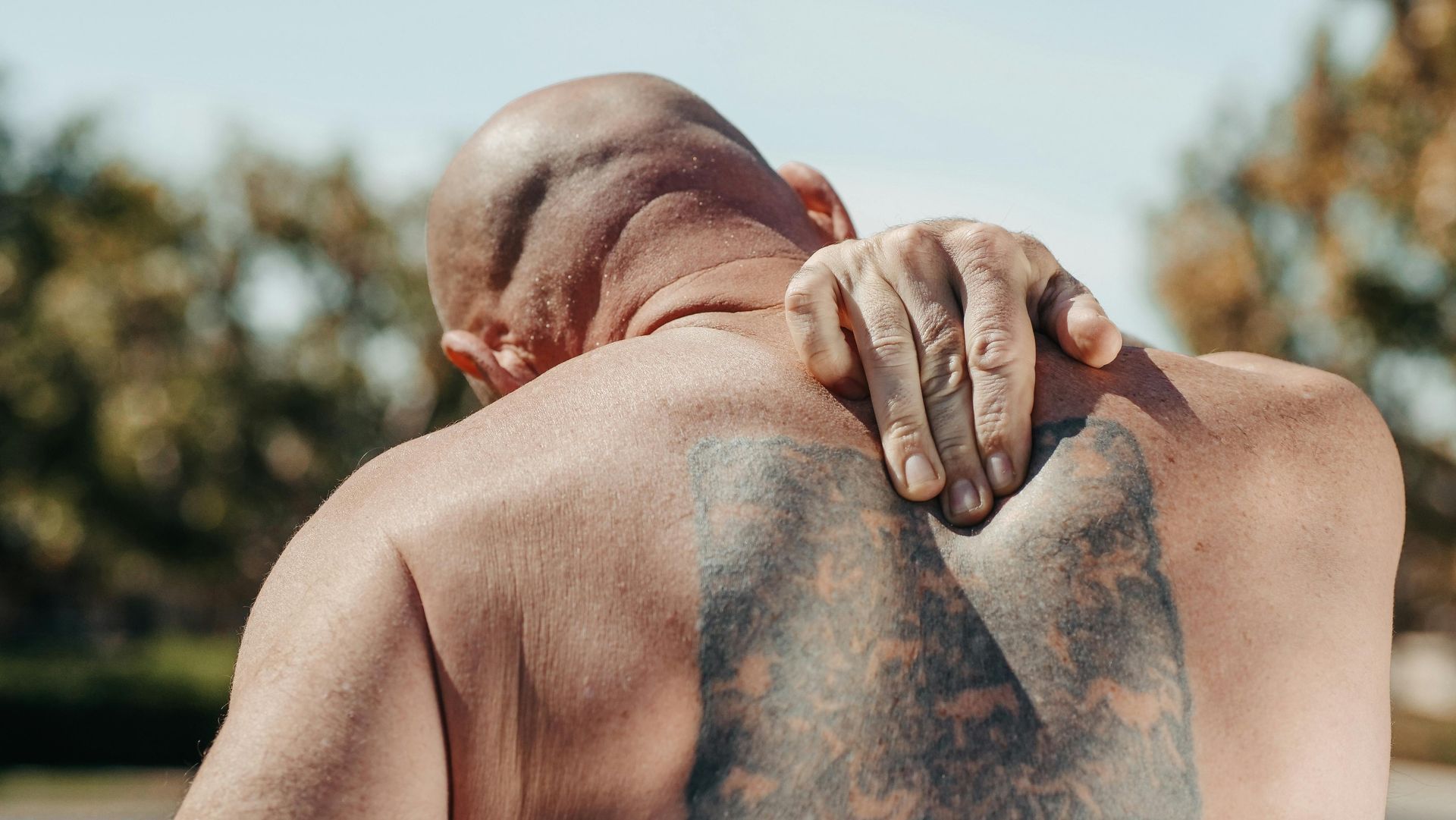 A man with a tattoo on his back is holding his shoulder in pain.