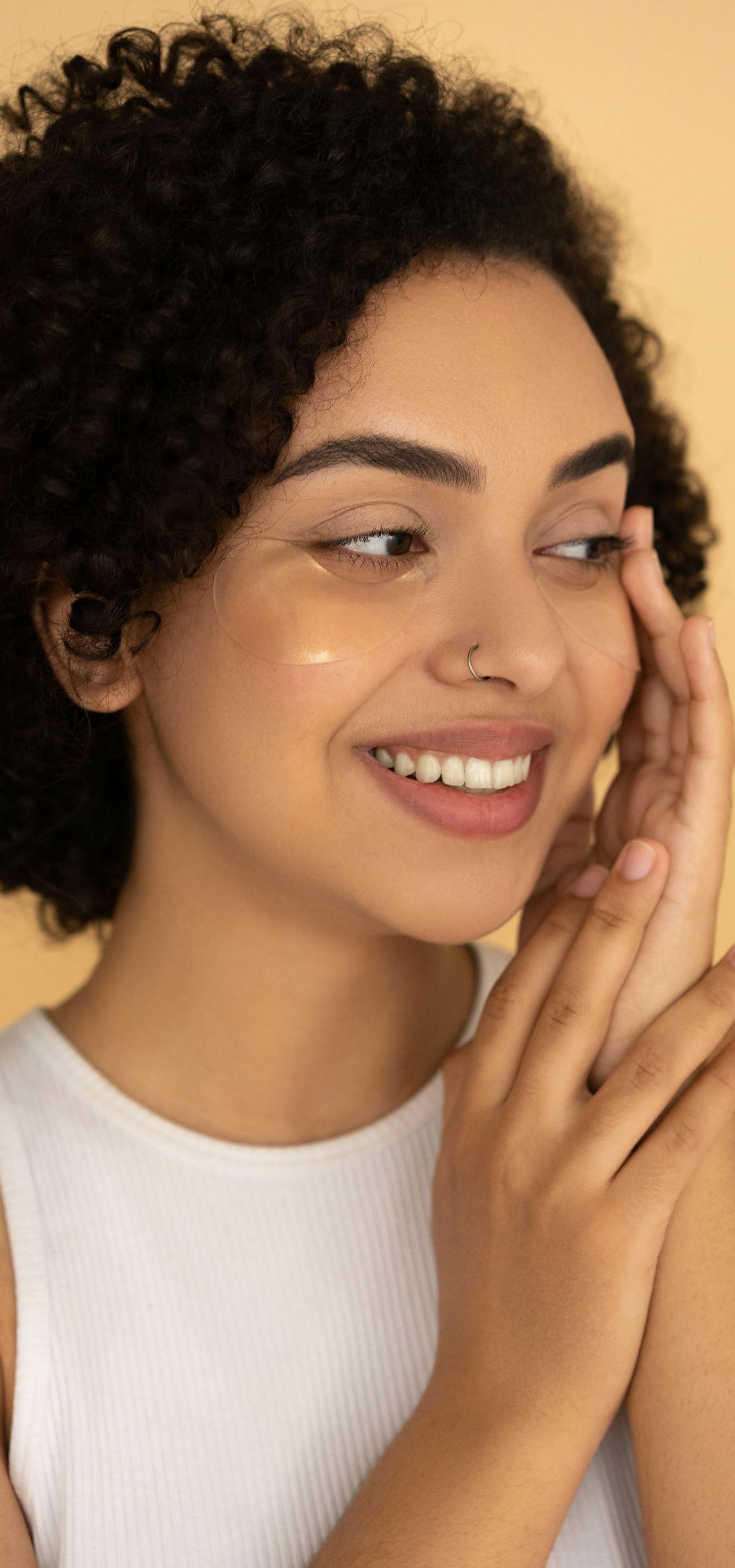 A woman with curly hair is smiling and touching her face.