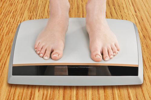 Feet standing on a digital scale, measuring weight. Light skin tones against a gray and black scale on a wood-grain floor.