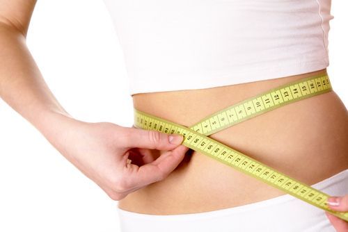 Woman in white top and underwear measuring waist with yellow tape measure.