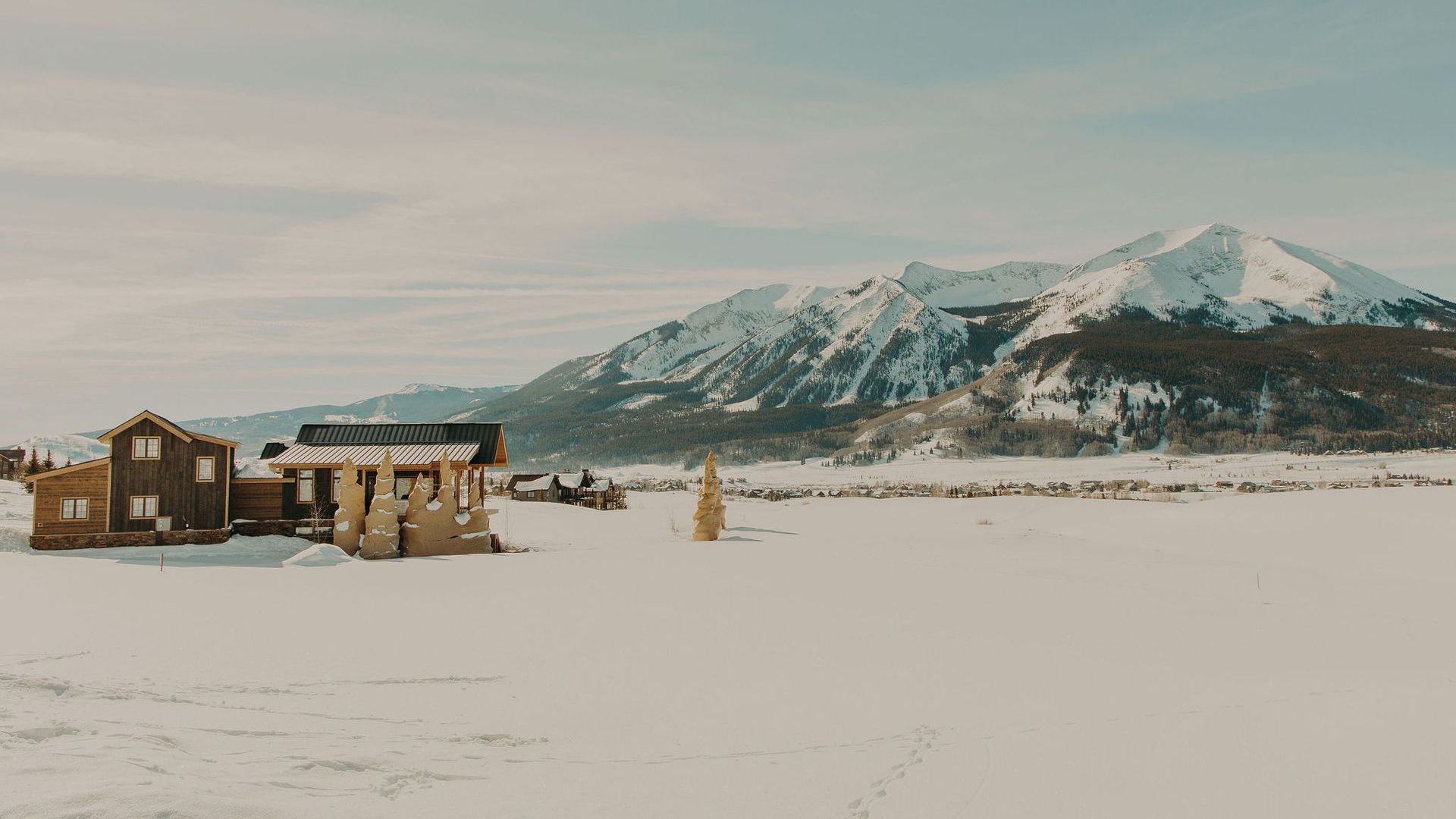 A couple of houses are sitting in the middle of a snow covered field with mountains in the background.
