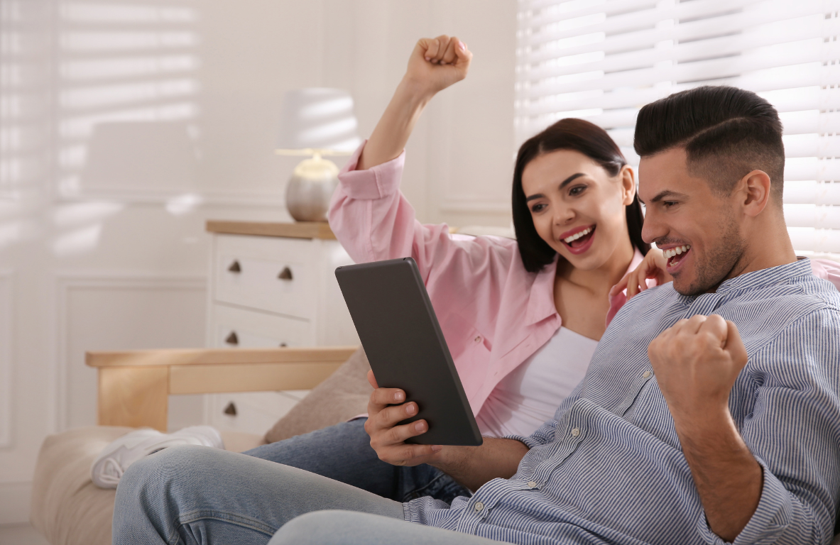 Two smiling people on a couch celebrating while looking at a tablet in a bright living room