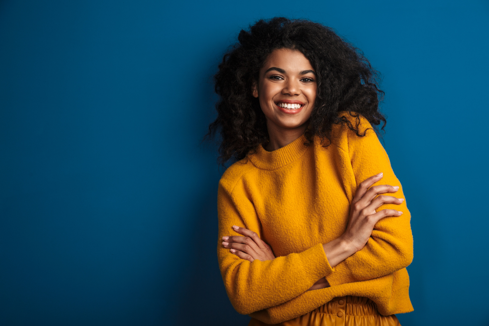 Smiling person in a mustard sweater with arms crossed against a blue background