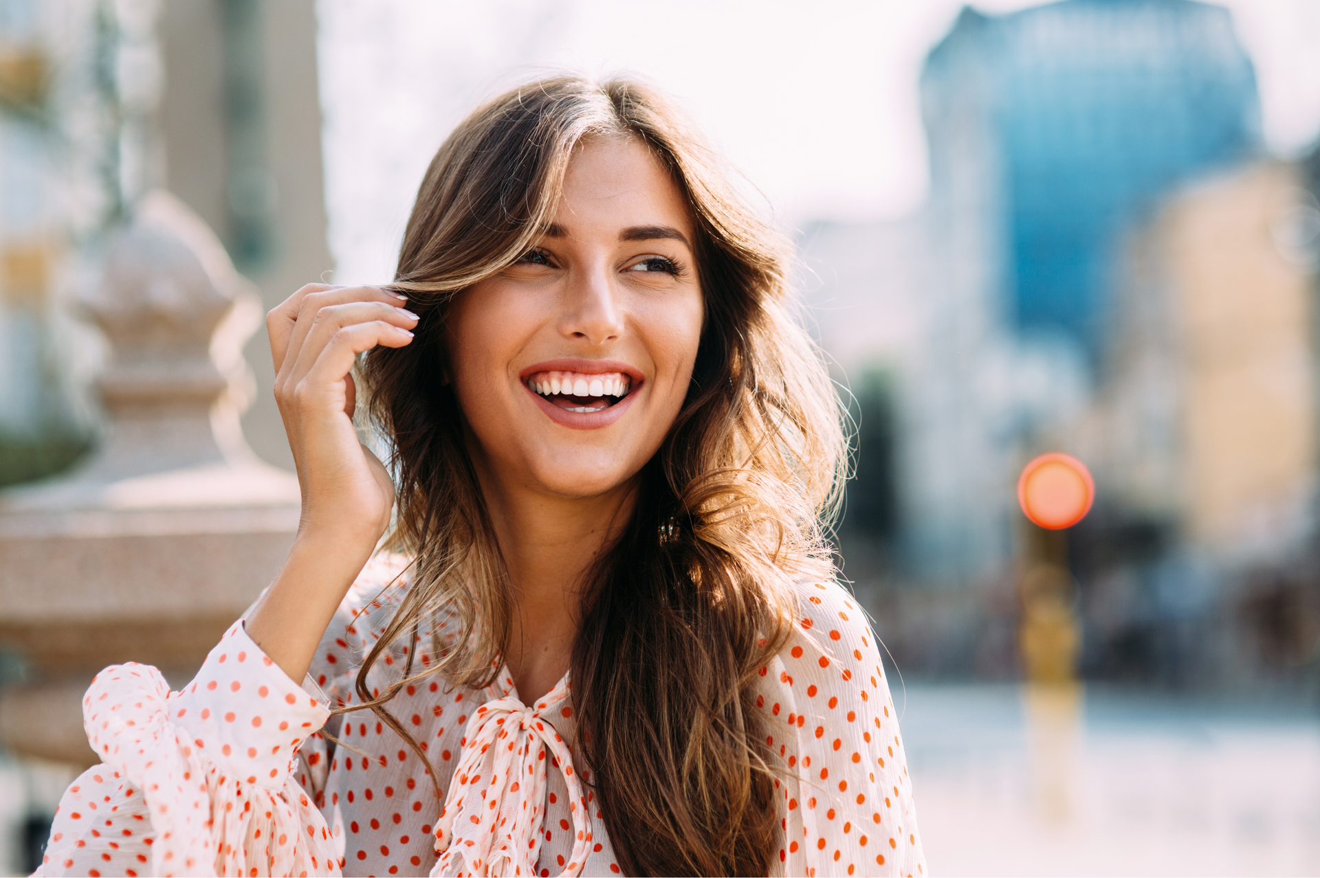 Smiling woman in a pink polka dot blouse outdoors, with a blurred city street behind her
