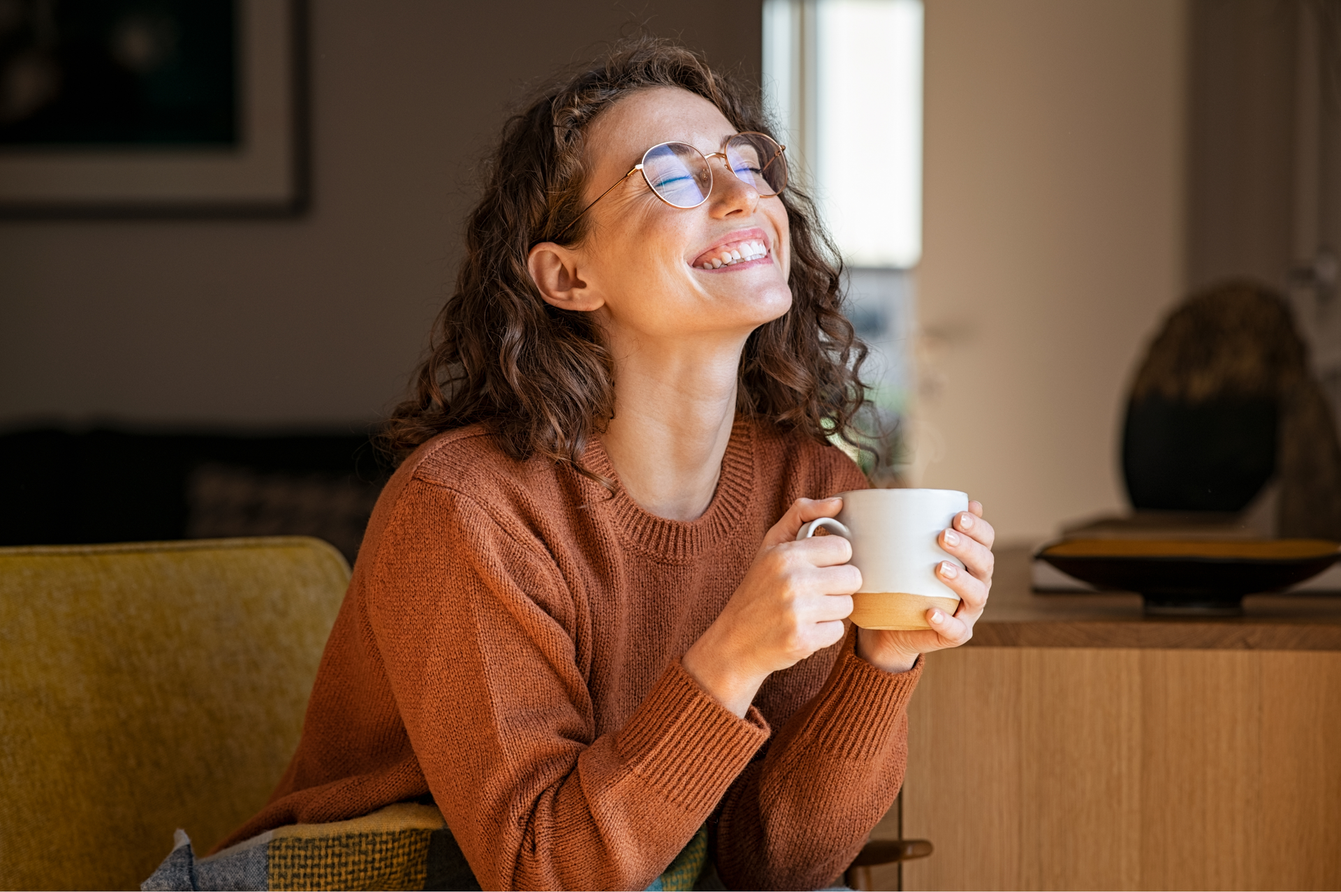 Smiling woman in a rust sweater holding a white mug on a couch indoors