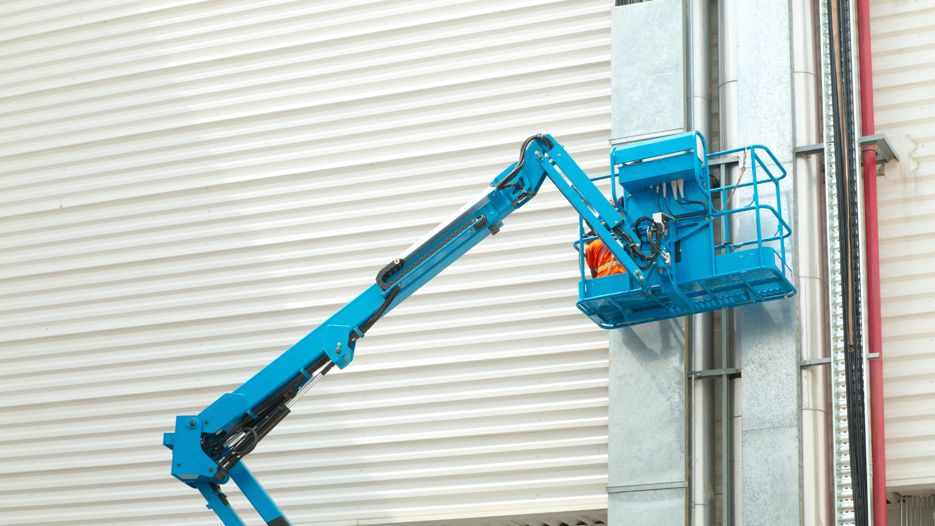 Blue articulated boom lift reaching towards a vertical metal structure, against a white wall.