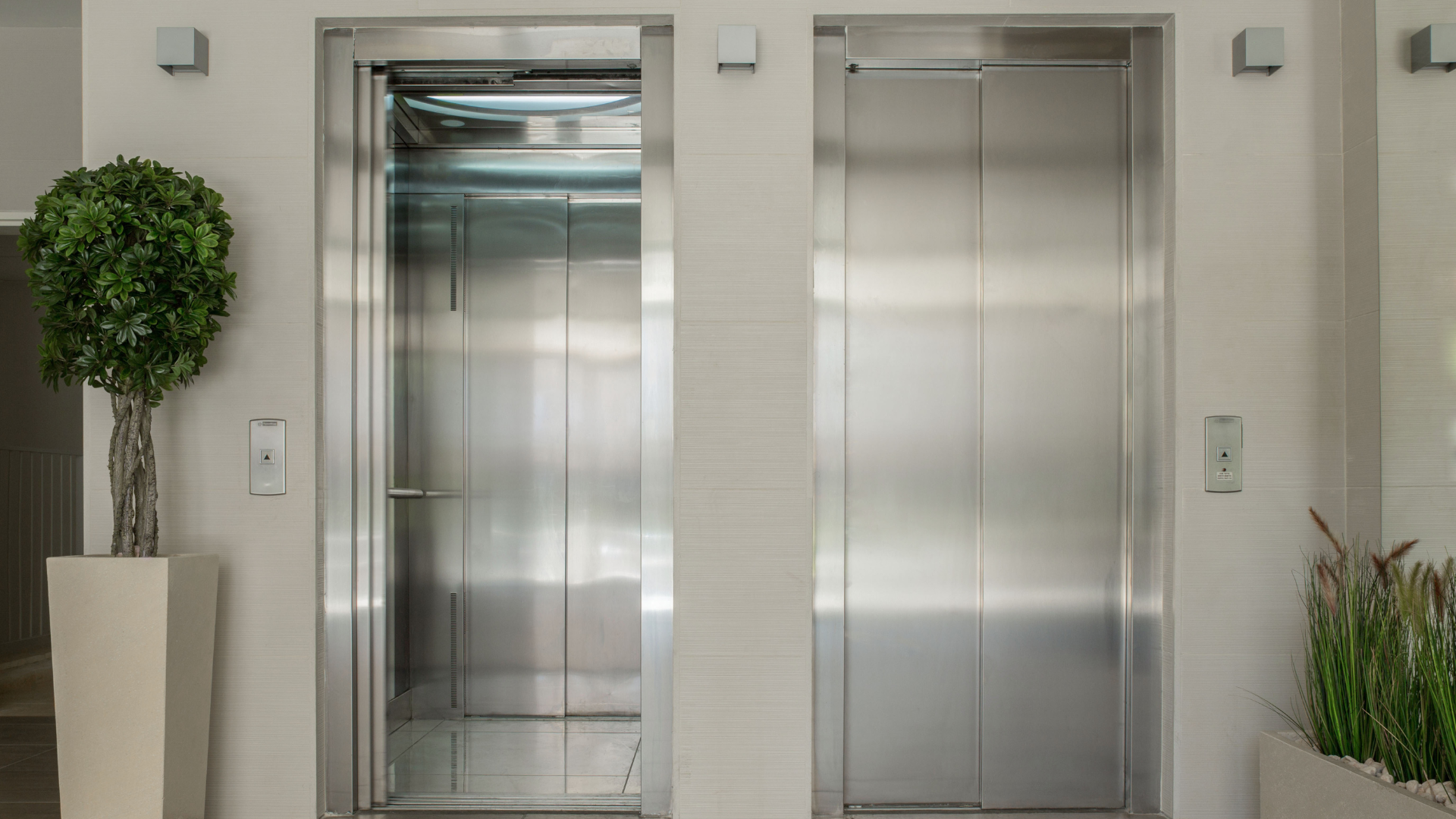 Two stainless steel elevators. One door open, one closed. White tiled wall. Potted greenery on either side.