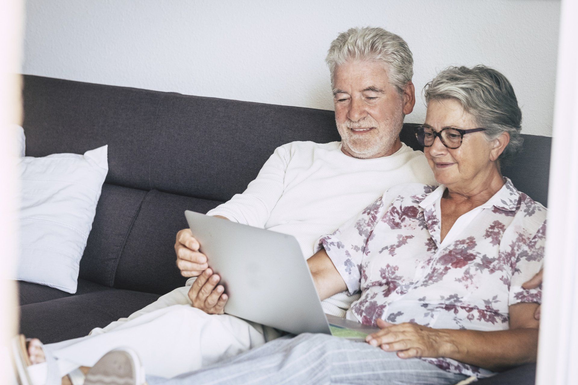 Elderly couple on sofa, looking at laptop, smiling.