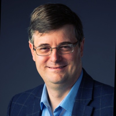 Man with glasses smiles, wearing blue shirt and plaid jacket, against a dark blue background.