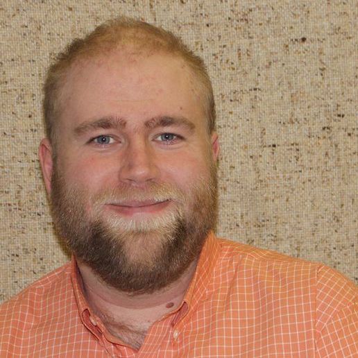 A man with a beard is wearing an orange shirt and smiling.