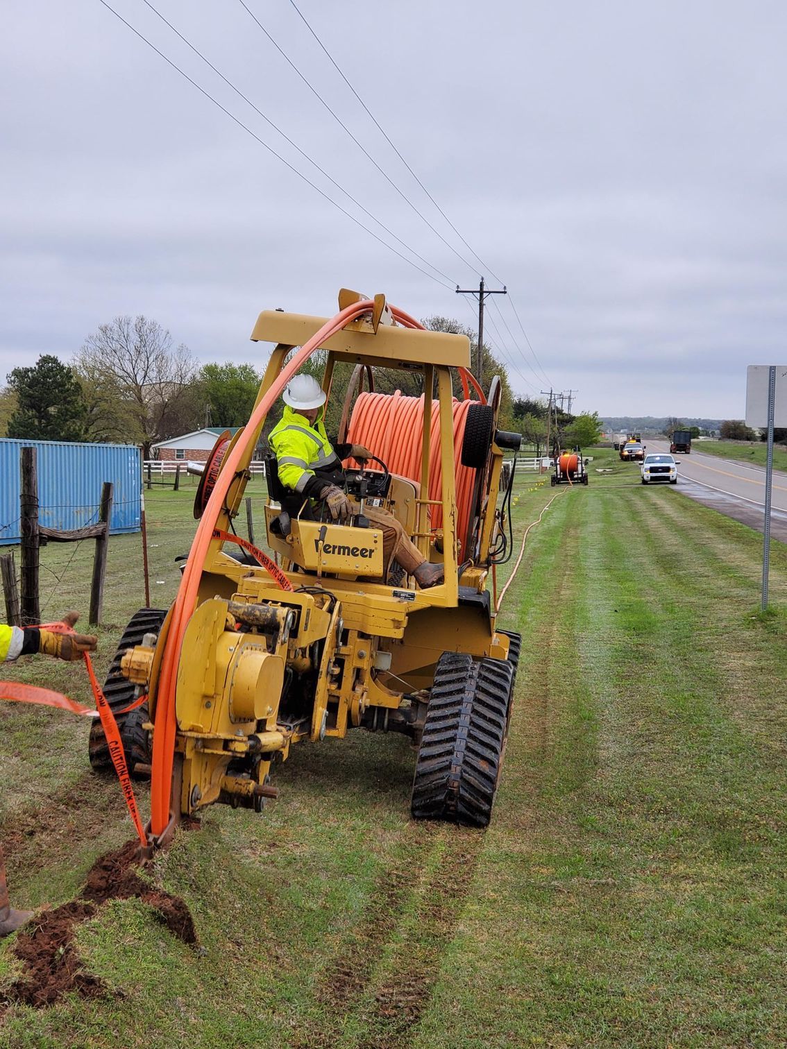A yellow trencher laying cable alongside a road. A worker operates the machine in an outdoor setting.