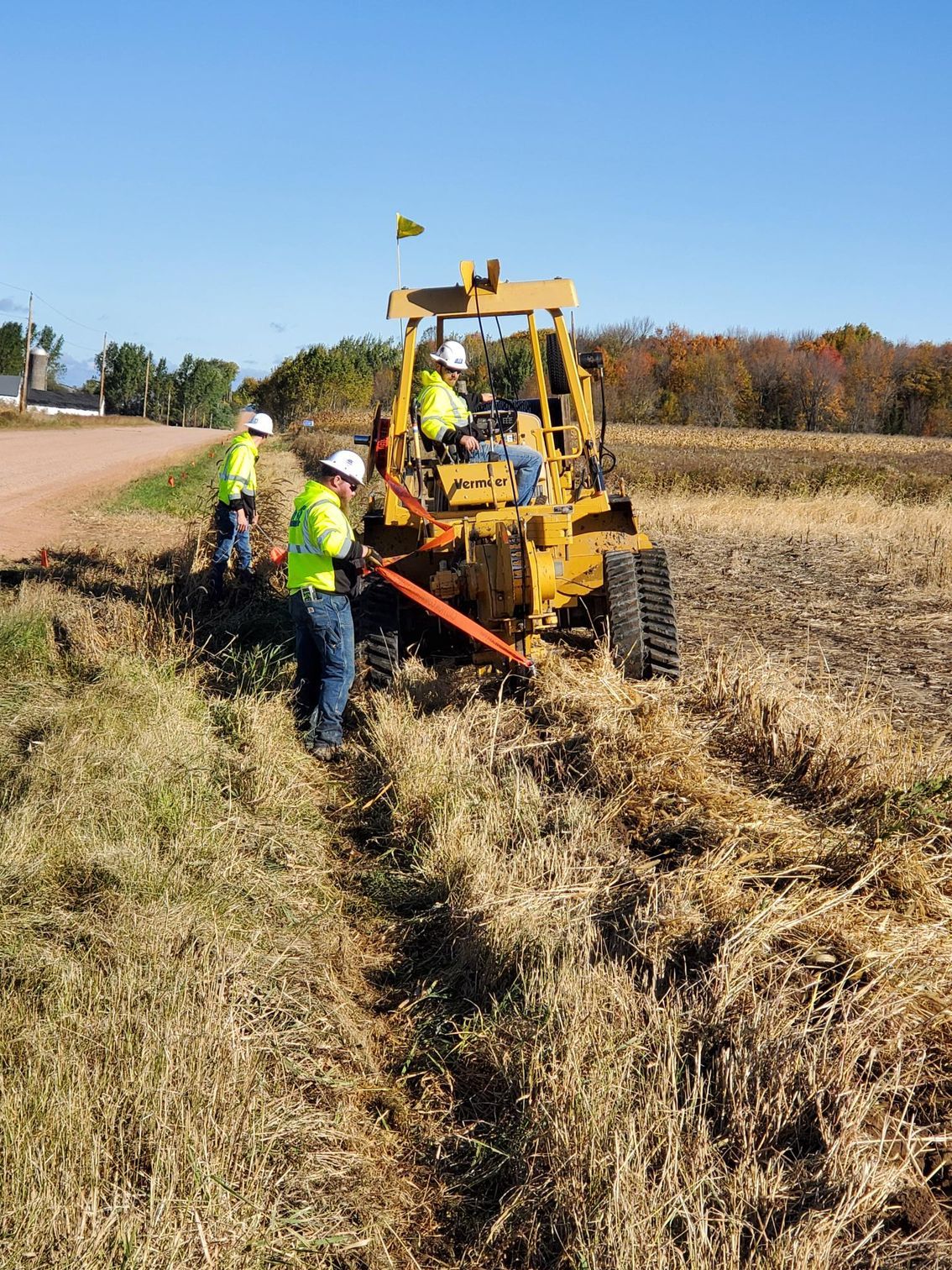 Workers operating a yellow trencher in a field. Two workers in safety vests and hard hats are working with the machine.