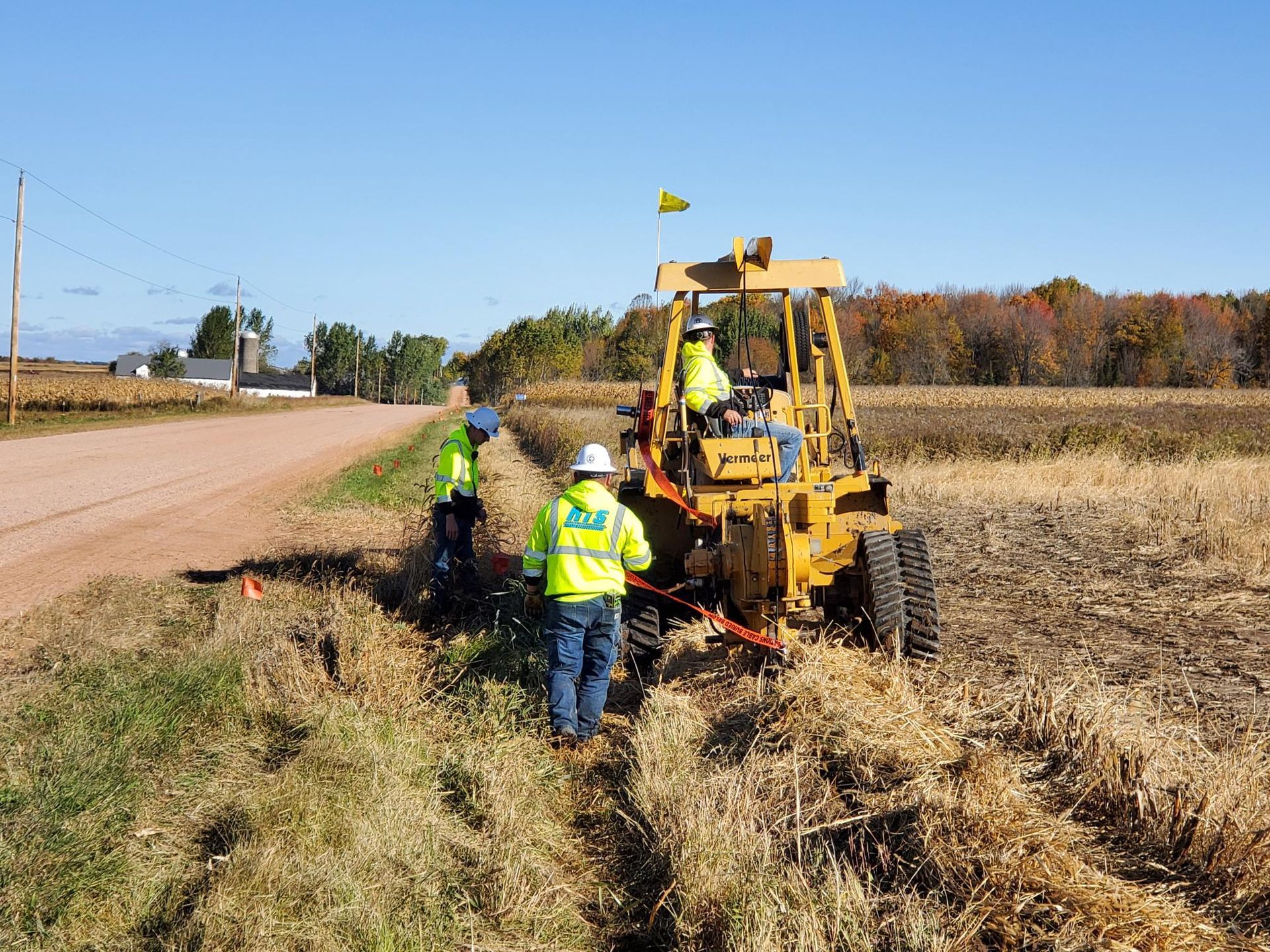 Workers in safety vests use a yellow trencher machine next to a road and a cornfield.