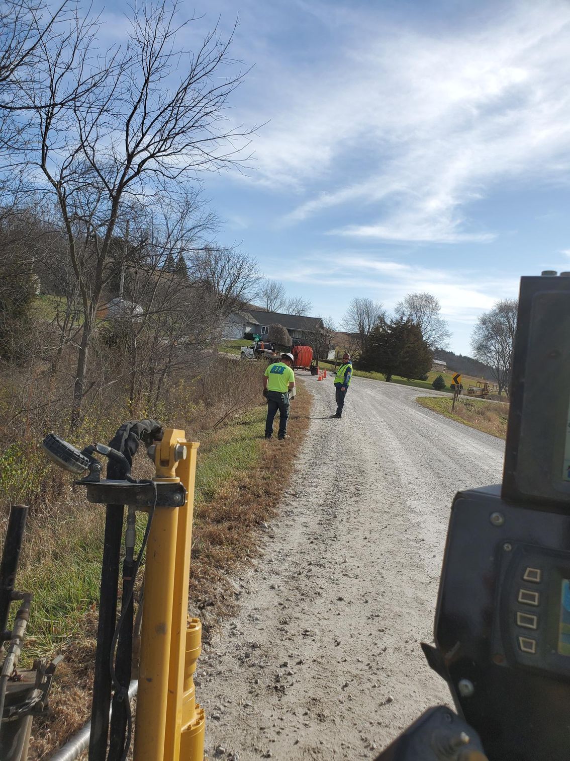 Two people in safety vests on a gravel road, near equipment, with trees and a house in the background.