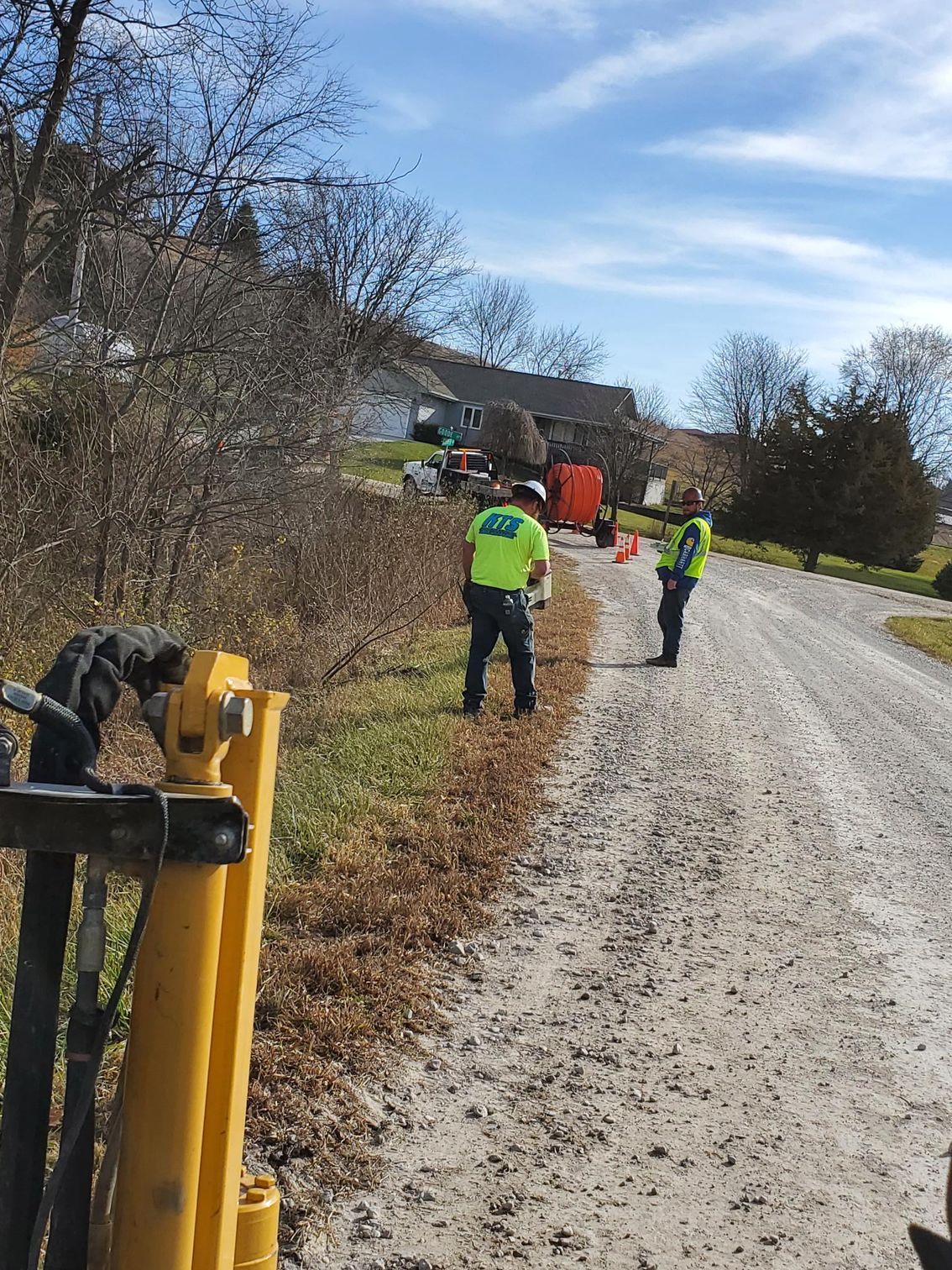 Two workers in safety vests stand near a gravel road, orange cones and equipment visible, with a house in the background.