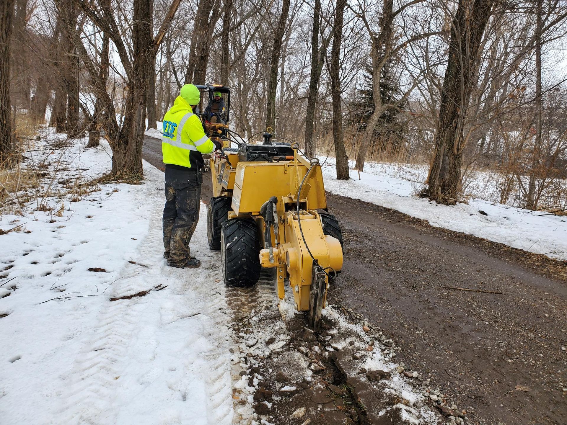 Person operating a yellow asphalt milling machine on a snow-covered road. Trees line the sides.