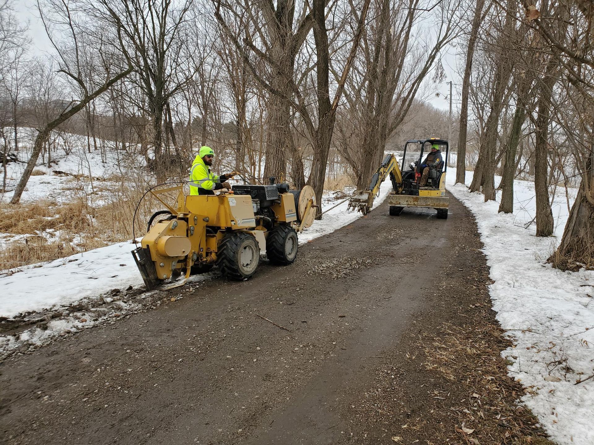 Two construction vehicles, removing debris from a path lined with bare trees and snow.