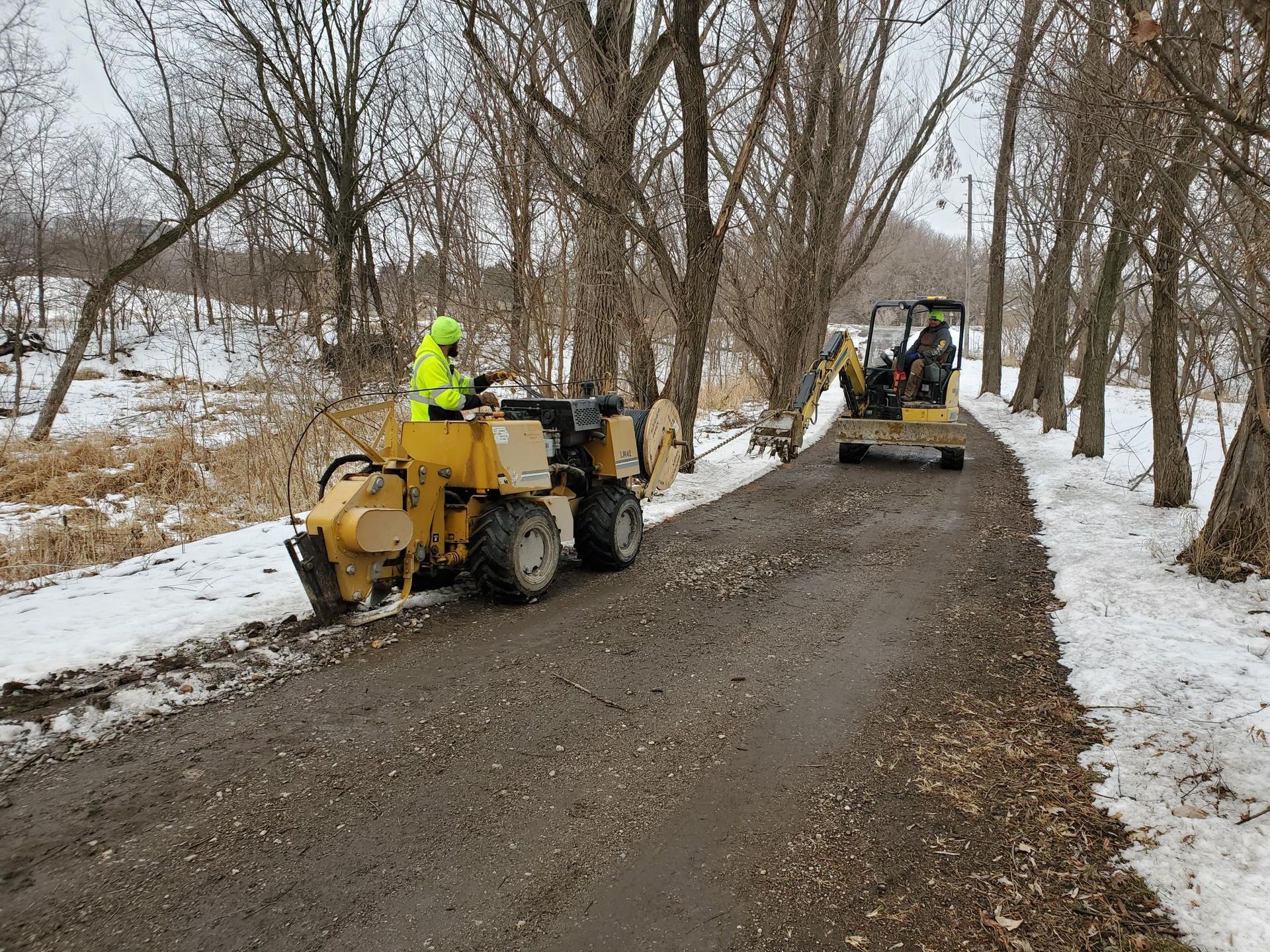 Construction equipment on a snowy road; an operator in a yellow safety vest grinds tree stumps, another drives an excavator.