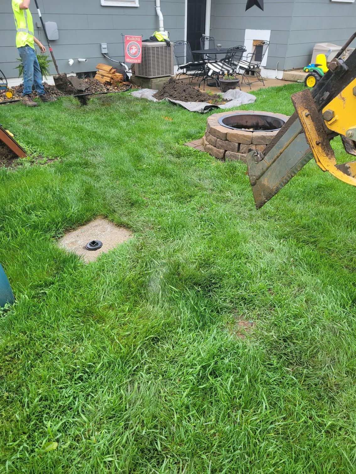 Yellow excavator working in a grassy backyard with a fire pit and a utility access cover.