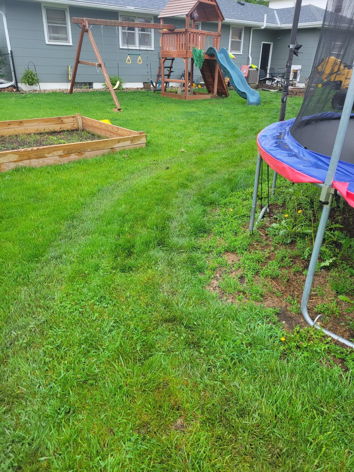 Backyard with green grass, swing set, garden bed, and a trampoline.